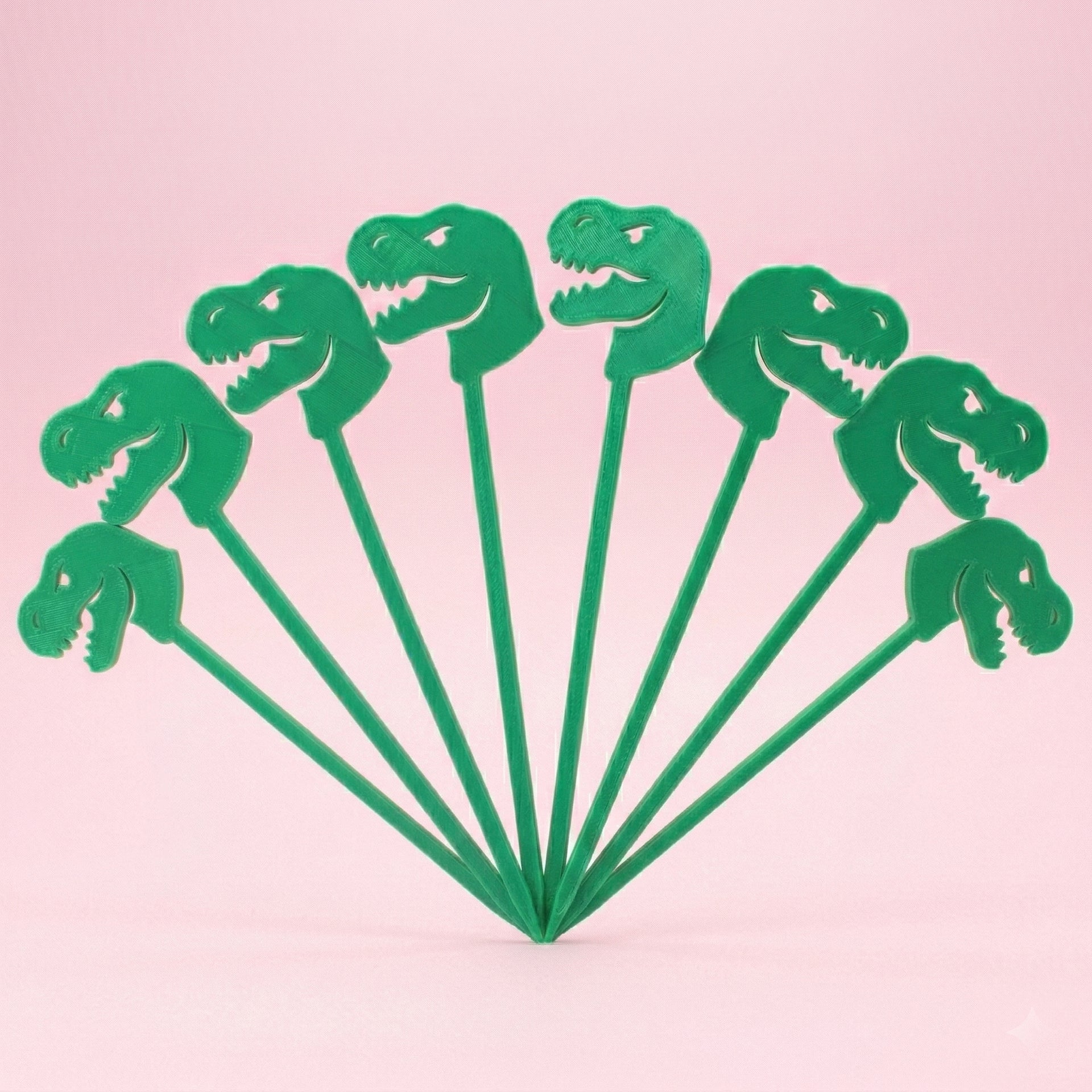 Huit piques apéritif en forme de tête de dinosaure T-Rex vert imprimés en 3D et disposés en éventail sur un fond rose. Accessoires originaux pour décoration de gâteau ou buffet d'anniversaire enfant sur le thème des dinosaures.