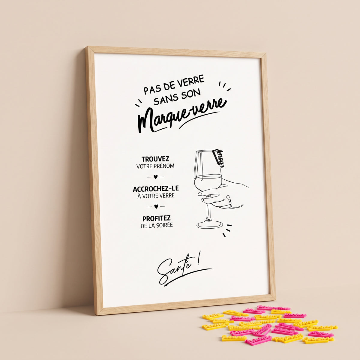 Grande affiche verticale « Pas de verre sans son marque-verre », imprimée sur un papier blanc, insérée dans un cadre en bois clair et appuyée contre un mur. Le tout est posé au sol sur un fond uni beige clair, pastel et lumineux. Les illustrations de l'affiche, avec une main qui tient un verre à pied et les consignes pour les invités, sont entièrement visibles. À la base du cadre, sur le sol à droite, un ensemble de tout petits marque-verres roses et dorés.