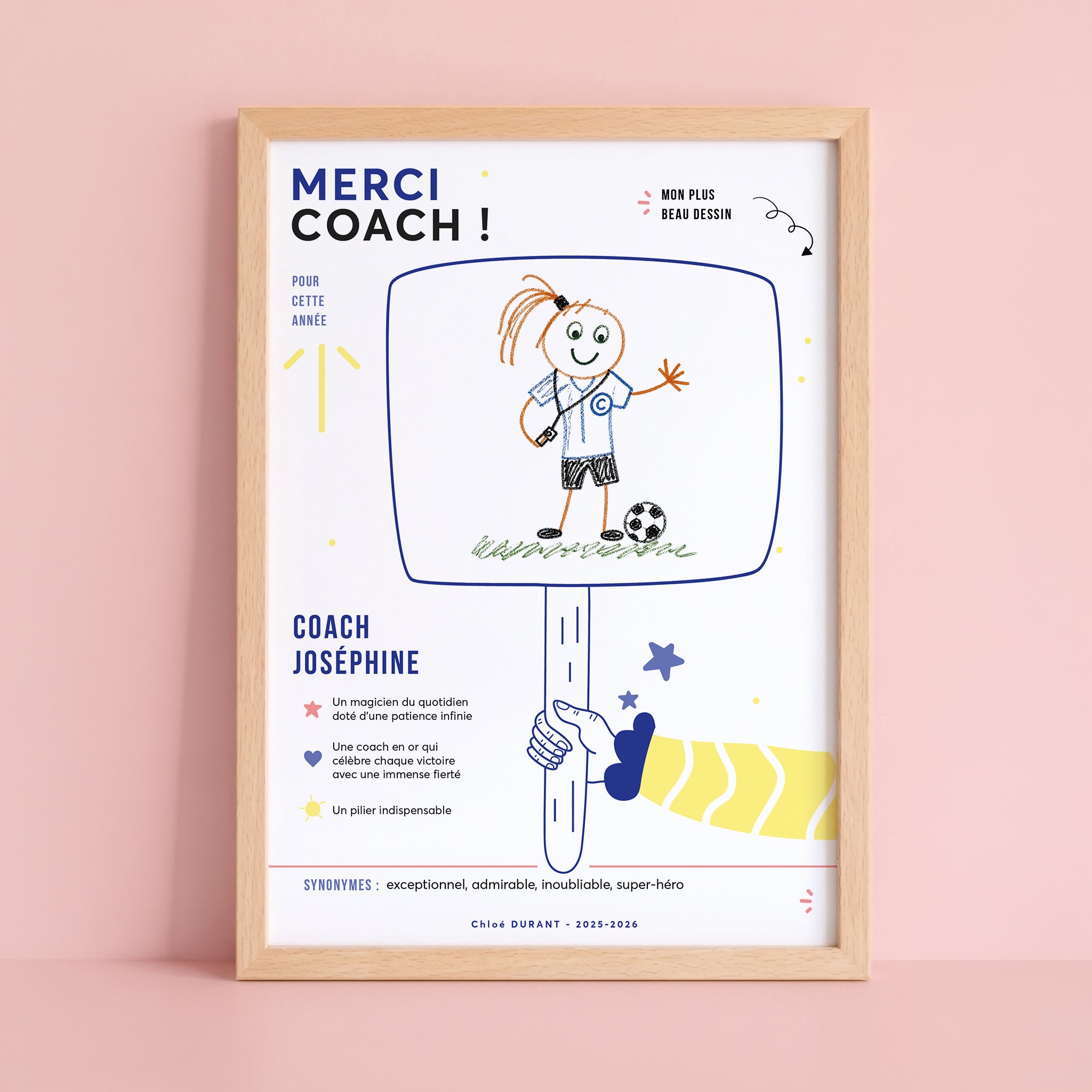 Une affiche de remerciement personnalisée, encadrée dans un bois clair et posée sur une surface rose pastel contre un mur rose. Le haut de l'affiche comporte le texte en gros caractères bleus "MERCI COACH !", accompagné de textes plus petits "POUR CETTE ANNÉE" et d'un texte manuscrit d'enfant "MON PLUS BEAU DESSIN". Le centre présente un dessin d'enfant au trait de crayon de couleur représentant une entraîneuse de football souriante en maillot bleu avec un "C" et un sifflet, debout sur de l'herbe avec un ba
