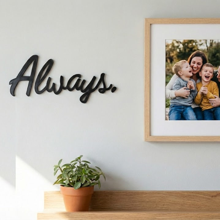Décoration murale “Always.” en impression 3D (PLA) noire, fixée sur un mur clair au-dessus d’une étagère en bois avec plante en pot, à côté d’un cadre photo.