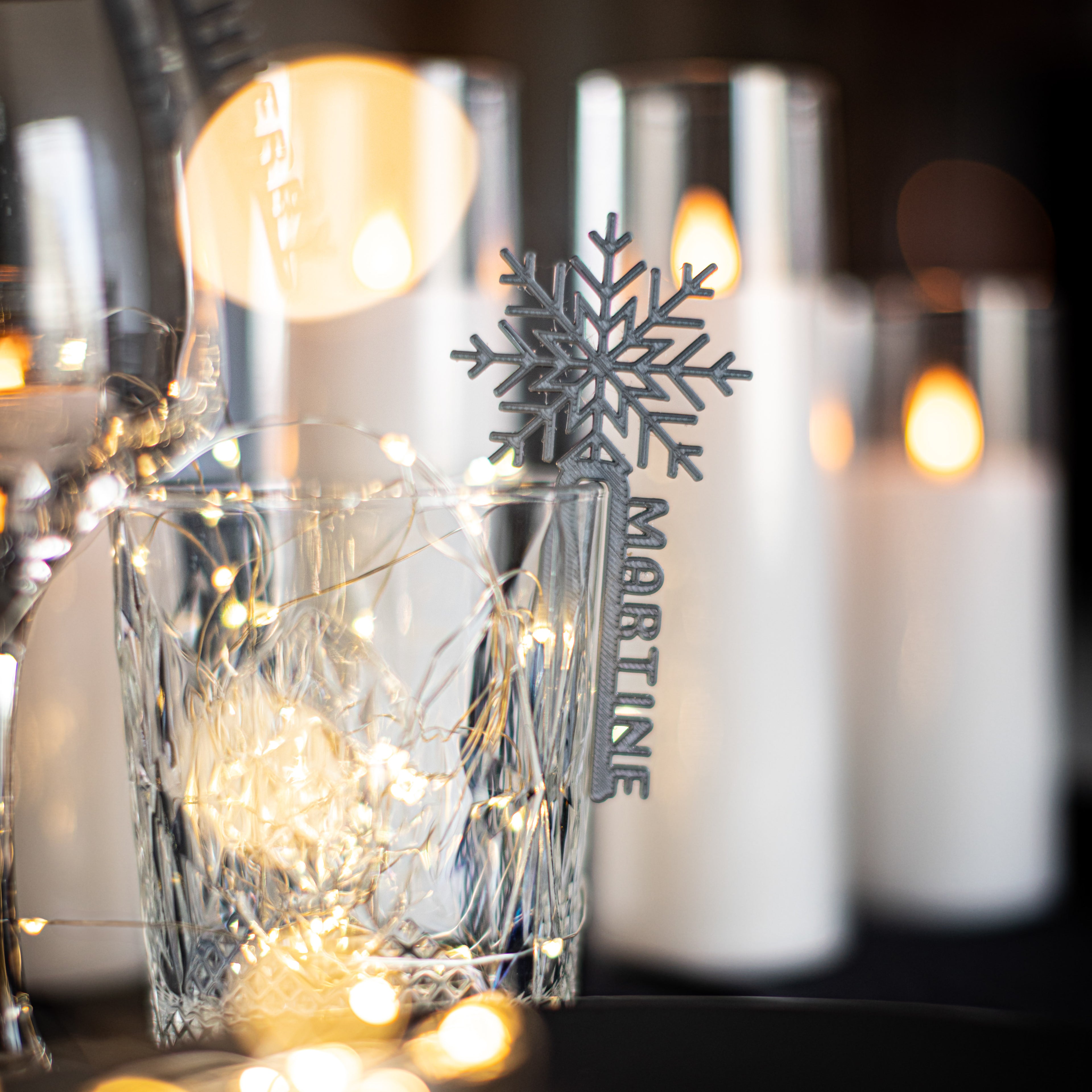 Marque-verre Flocon argenté personnalisé “Martine” accroché à un verre transparent, entouré de guirlandes lumineuses et de bougies LED pour une décoration d’hiver chaleureuse et élégante.