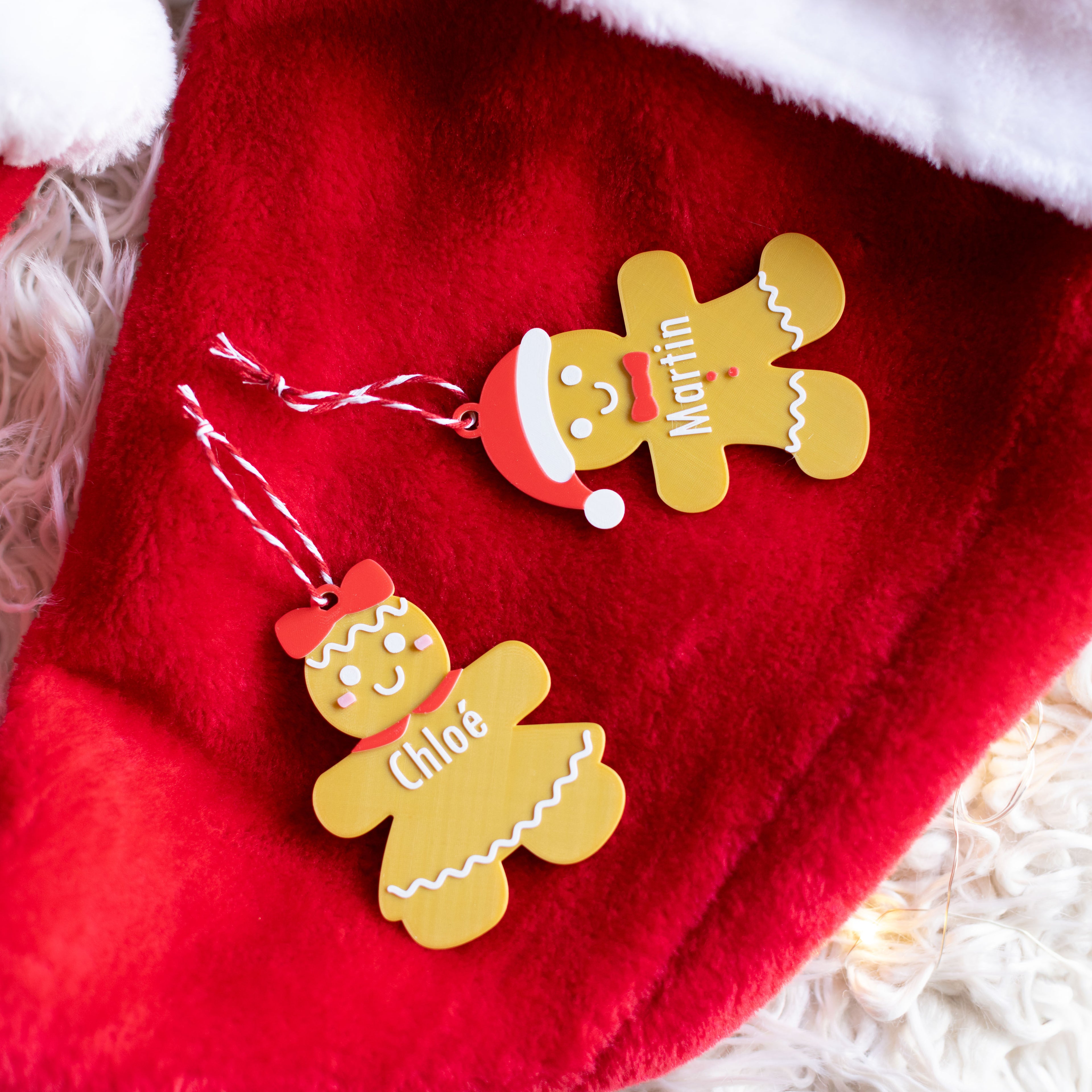 Deux décorations de Noël personnalisables en forme de bonhomme en pain d’épice : un personnage avec bonnet de Père Noël (prénom “Martin”) et une avec nœud (prénom “Chloé”), posés sur un tissu rouge avec ficelles de suspension rouge et blanc.