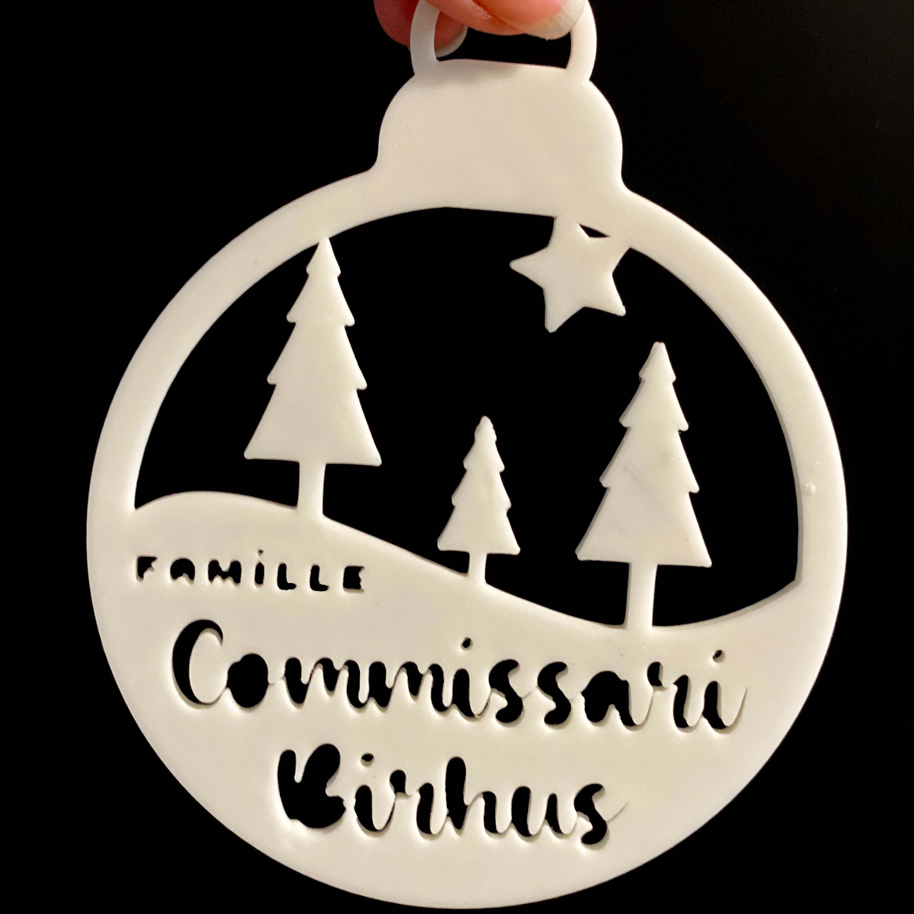 Gros plan d’une boule de Noël personnalisable “La forêt familiale” : décoration blanche découpée avec trois sapins et une étoile, texte “Famille” + nom/prénom (ex. “Commissaire Birhus”), suspendue sur fond noir.