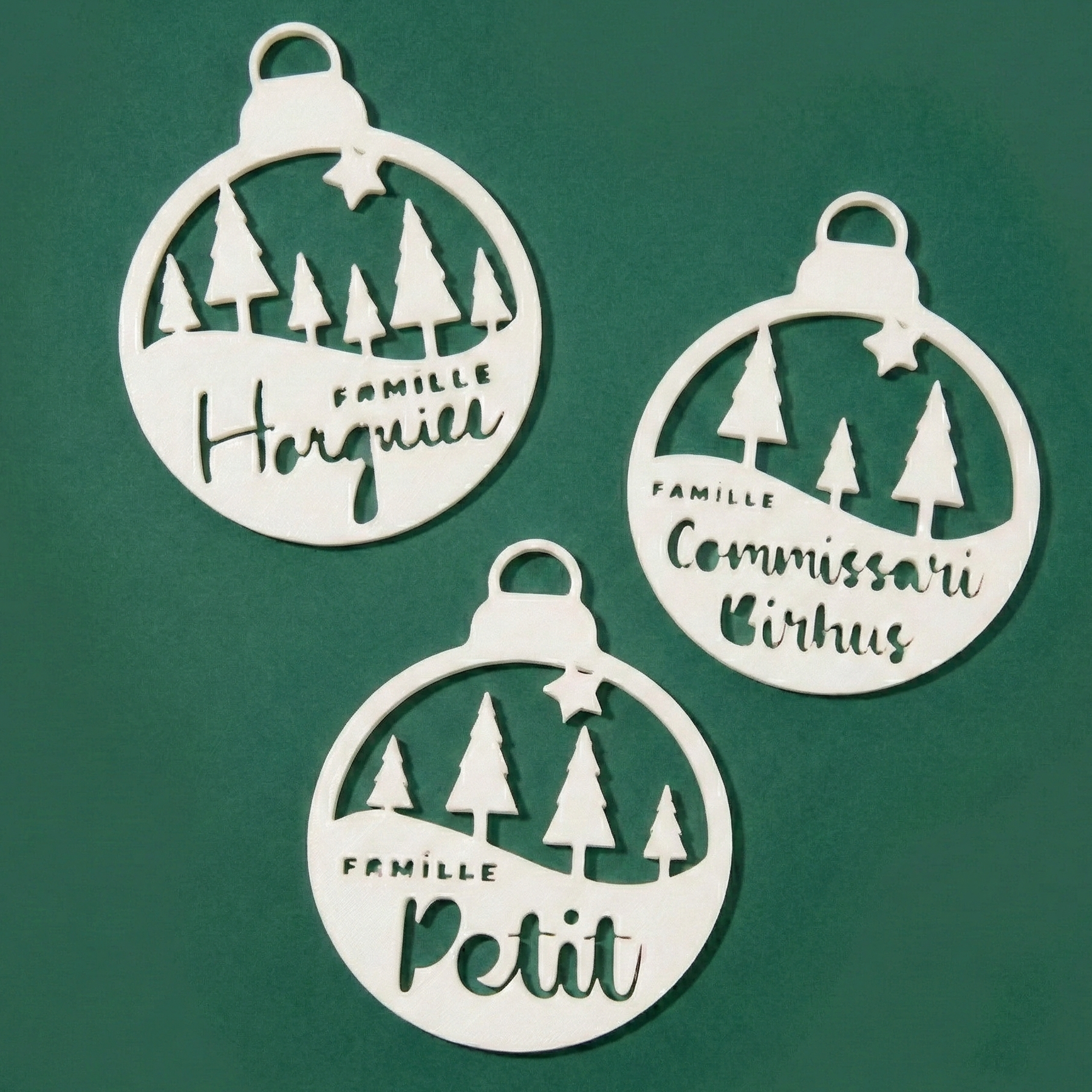 Trois boules de Noël personnalisables “La forêt familiale” : décor découpé avec sapins et petite étoile, texte “Famille” + nom/prénom (ex. “Horgnies”, “Commissaire Birhus”, “Petit”), présentées sur fond vert.