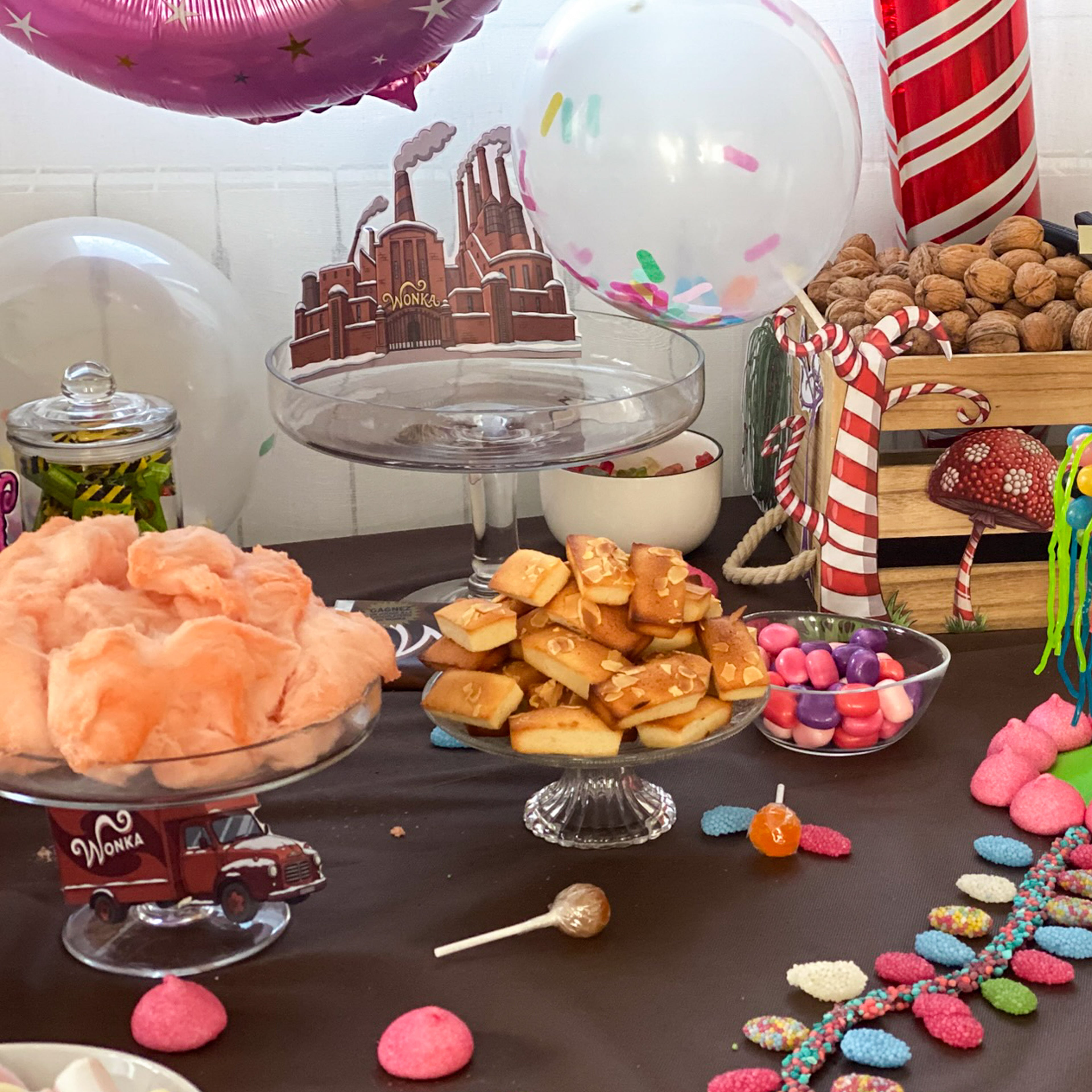 Décoration de table d'anniversaire sur le thème "Charlie et la chocolaterie", comprenant des plateaux de barbe à papa rose, des financiers, des bonbons colorés et des noix. Des illustrations de l'usine Wonka, un camion décoratif et des ballons festifs complètent cette mise en scène gourmande.