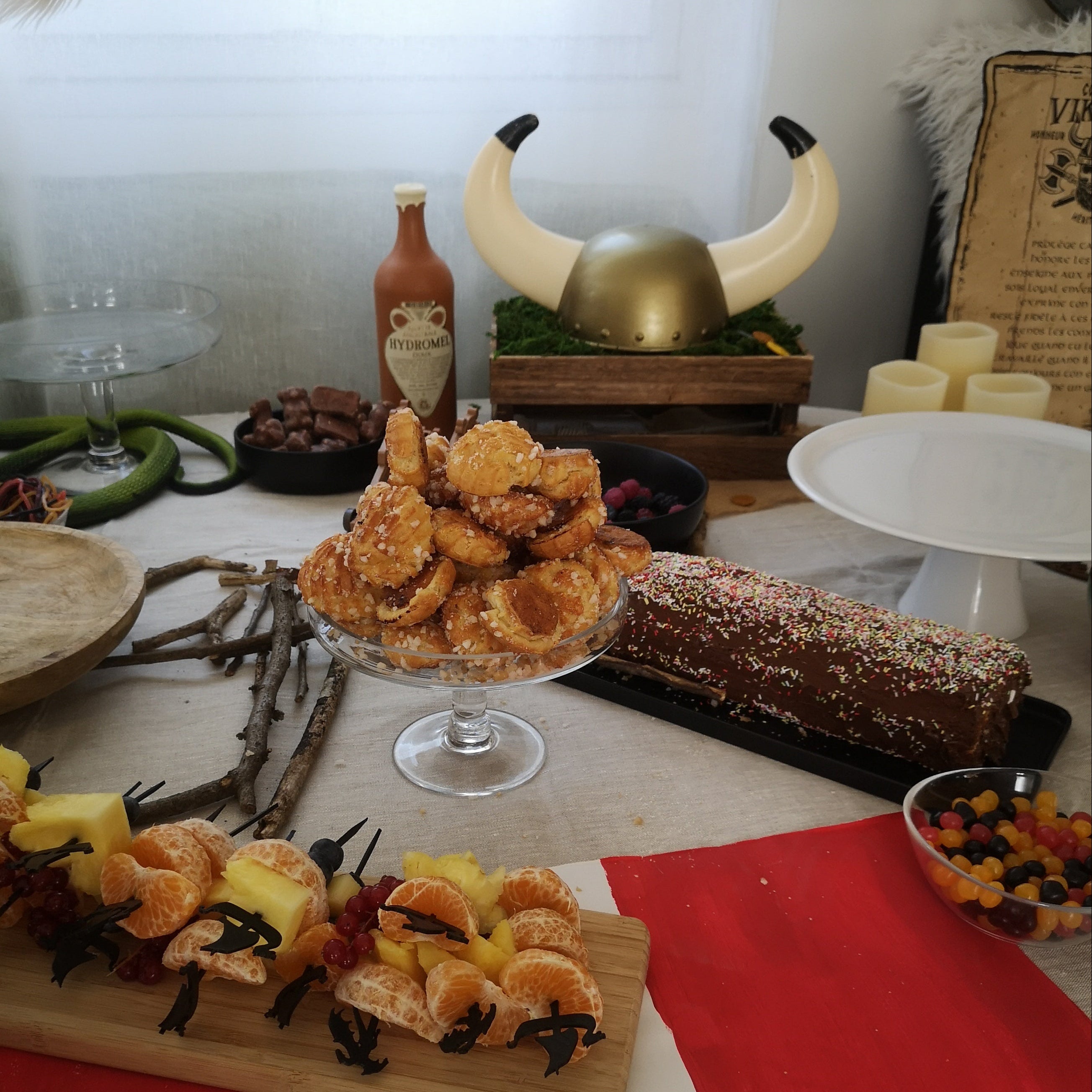 Mise en scène d'un buffet thématique scandinave ou viking mettant en avant des piques gourmet Drakkar en impression 3D noire. Au premier plan, un plateau en bois présente des mini-brochettes de fruits frais (clémentines, ananas et myrtilles) réalisées avec ces piques d'environ 14 cm de long. L'arrière-plan complète l'ambiance avec divers desserts, une décoration rustique à base de branches et un tissu rouge et blanc évoquant une voile de drakkar.