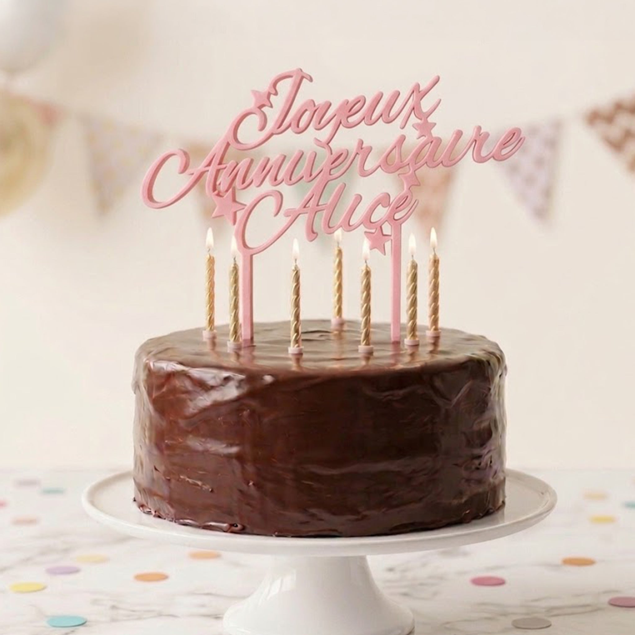 Cake topper personnalisé en script rose lisant 'Joyeux Anniversaire Alice', orné de petites étoiles, posé sur un gâteau au chocolat richement glacé avec six bougies dorées allumées. En arrière-plan, une scène de fête floue avec des ballons pastel et des fanions.