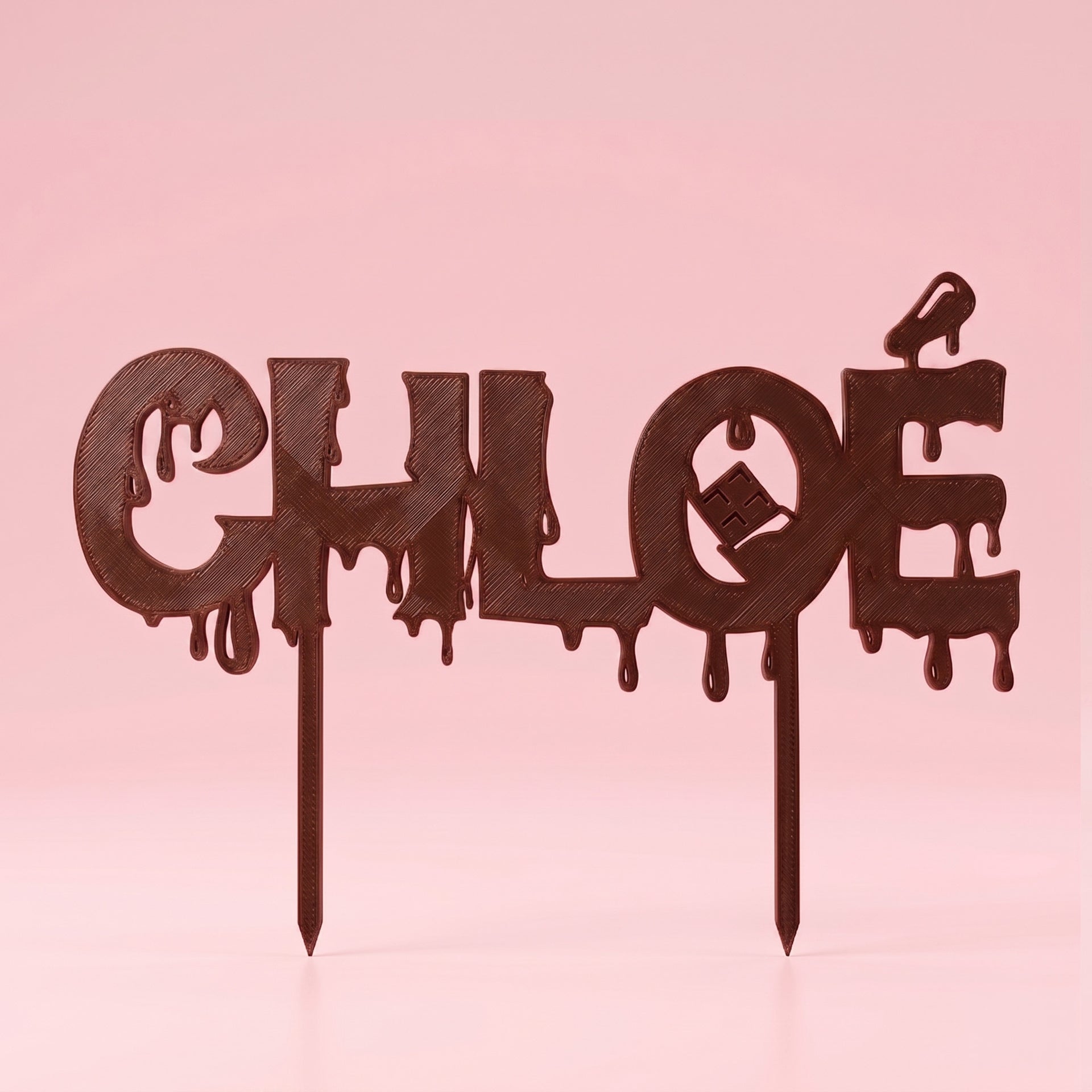 Packshot de face d'un cake topper personnalisé au prénom CHLOÉ, d'un coloris marron chocolat foncé et profond. L'objet présente un style de chocolat fondant avec des coulures et des détails d'impression 3D texturés (stries visibles). Un petit motif de tablette de chocolat est intégré au centre de la lettre 'O'. Porté par deux fines tiges pointues à sa base, le topper est centré au format carré sur un fond uni rose pastel doux et lumineux.