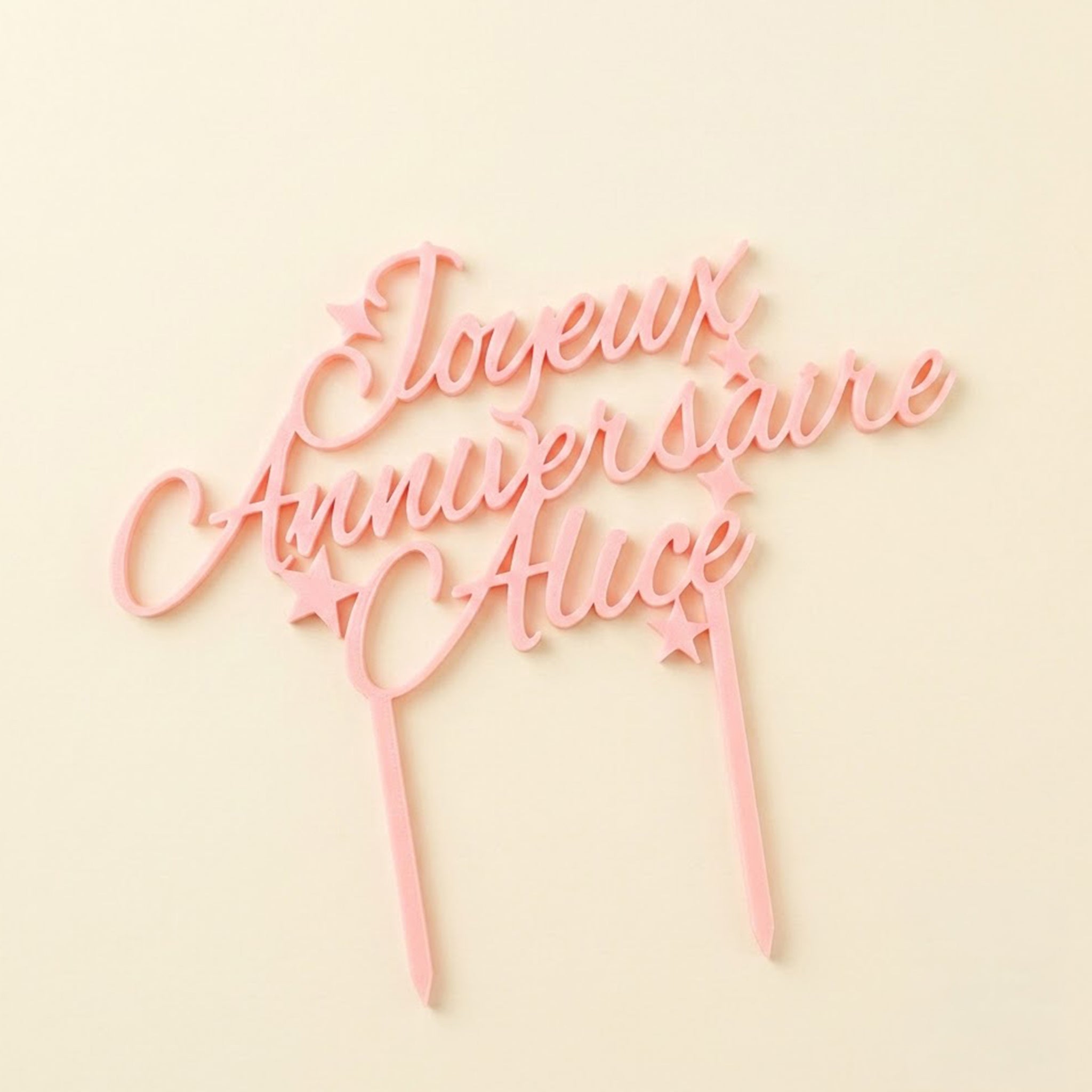 Cake topper personnalisé rose avec l'inscription cursive 'Joyeux Anniversaire Alice' et des petites étoiles, présenté à plat sur un fond uni jaune pastel.