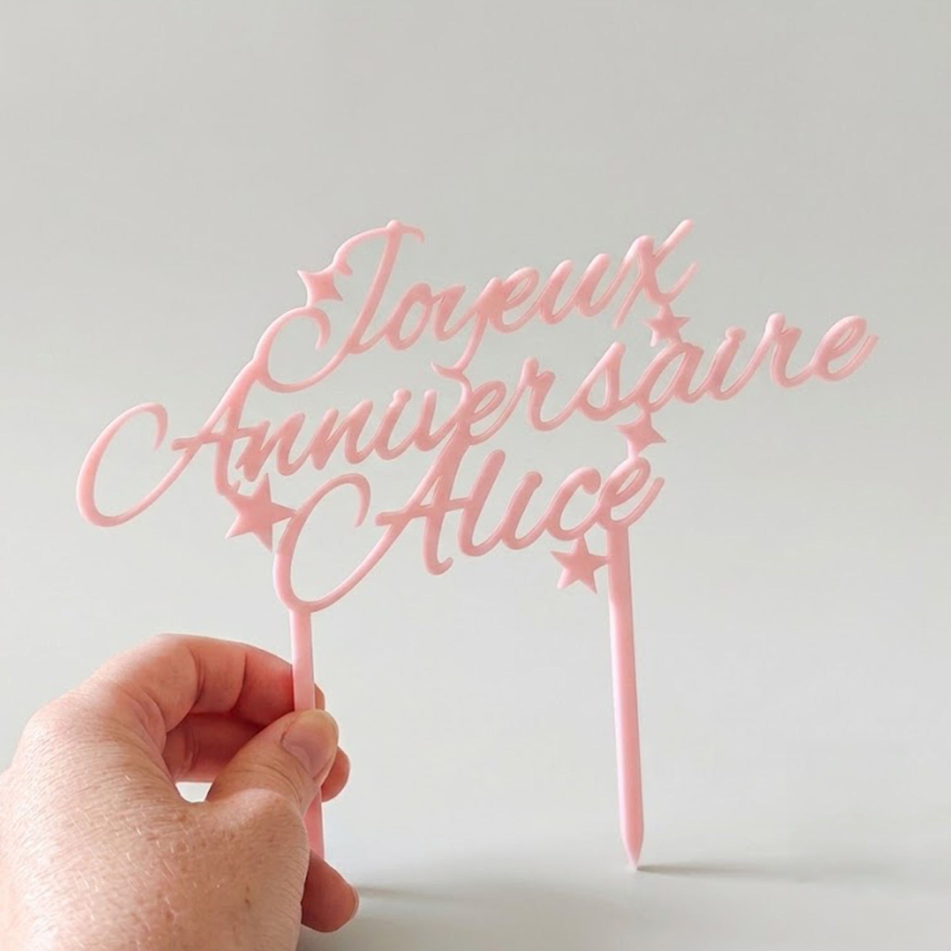 Main tenant un cake topper personnalisé en plastique rose avec l'inscription cursive 'Joyeux Anniversaire Alice' et des motifs de petites étoiles, sur un fond neutre épuré.