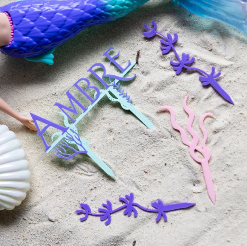 Lot de cake toppers sirène personnalisés avec le prénom Ambre, accompagné de décorations algues roses et violettes, posés sur du sable pour une décoration de gâteau d’anniversaire sur le thème des sirènes et du fond marin.