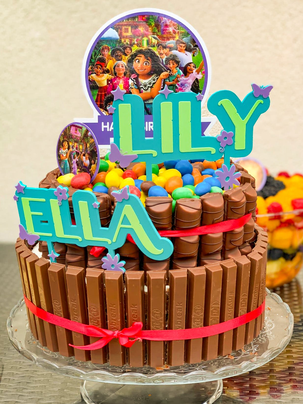 Cake topper personnalisé Encanto avec prénoms Lily et Ella sur gâteau d’anniversaire coloré, décoration festive inspirée de l’univers magique d’Encanto pour enfant.