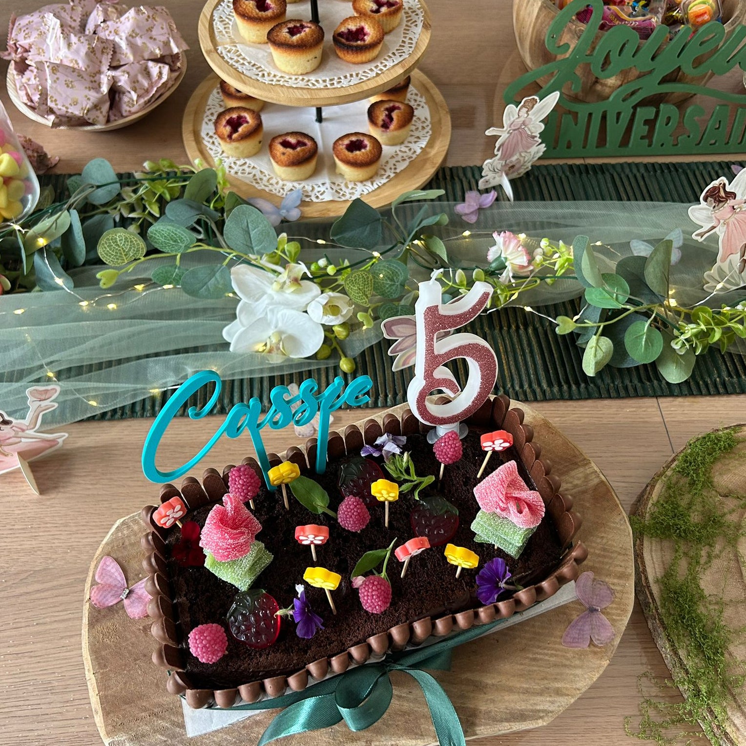Cake topper personnalisable prénom Élégance en bleu turquoise avec l'inscription "Cassie", posé sur un gâteau d'anniversaire décoré de framboises et de bonbons. Décoration de table sur le thème des fées en arrière-plan.