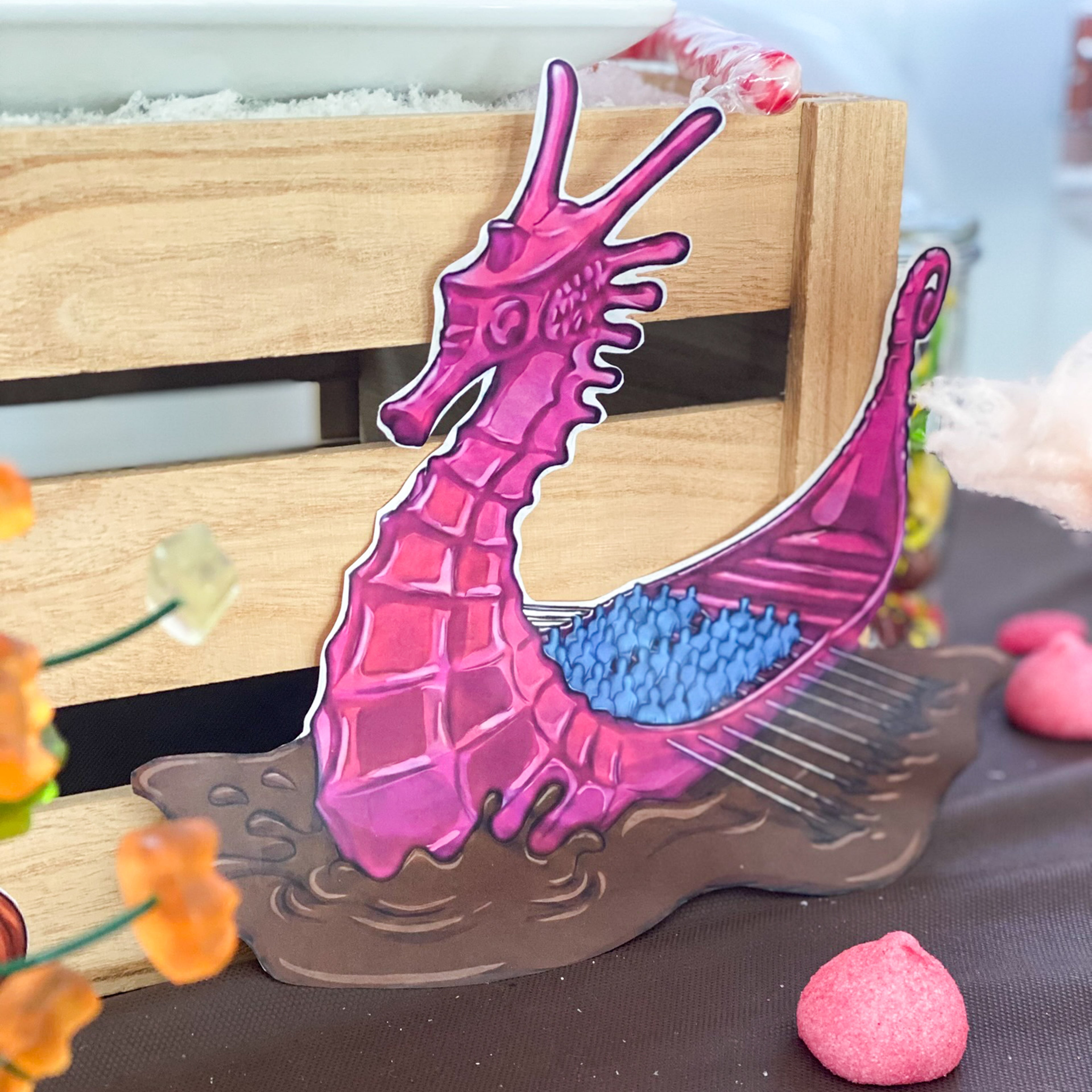 Gros plan sur l'illustration découpée du célèbre bateau rose en forme de dragon naviguant sur une rivière de chocolat. Placée contre une caisse en bois au milieu de bonbons, cette décoration en papier issue du kit PDF permet de recréer l'univers magique de la chocolaterie.