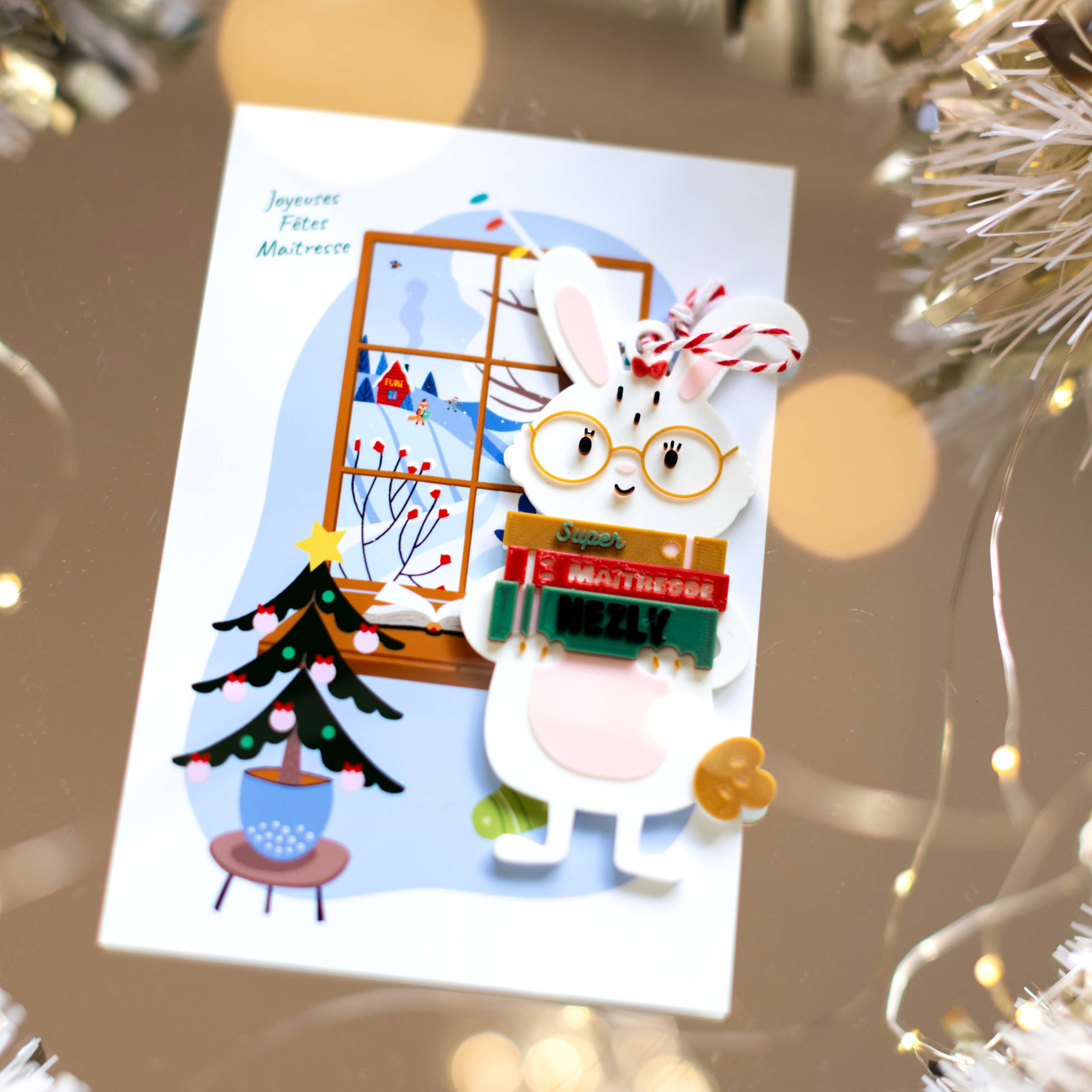 Carte “Joyeuses fêtes Maîtresse” avec une suspension d'un lapin décoratif portant des lunettes, tenant des pancartes “Super”, “Maîtresse” et un prénom personnalisé, devant une illustration de fenêtre d’hiver et sapin de Noël, avec ficelle rouge et blanche.