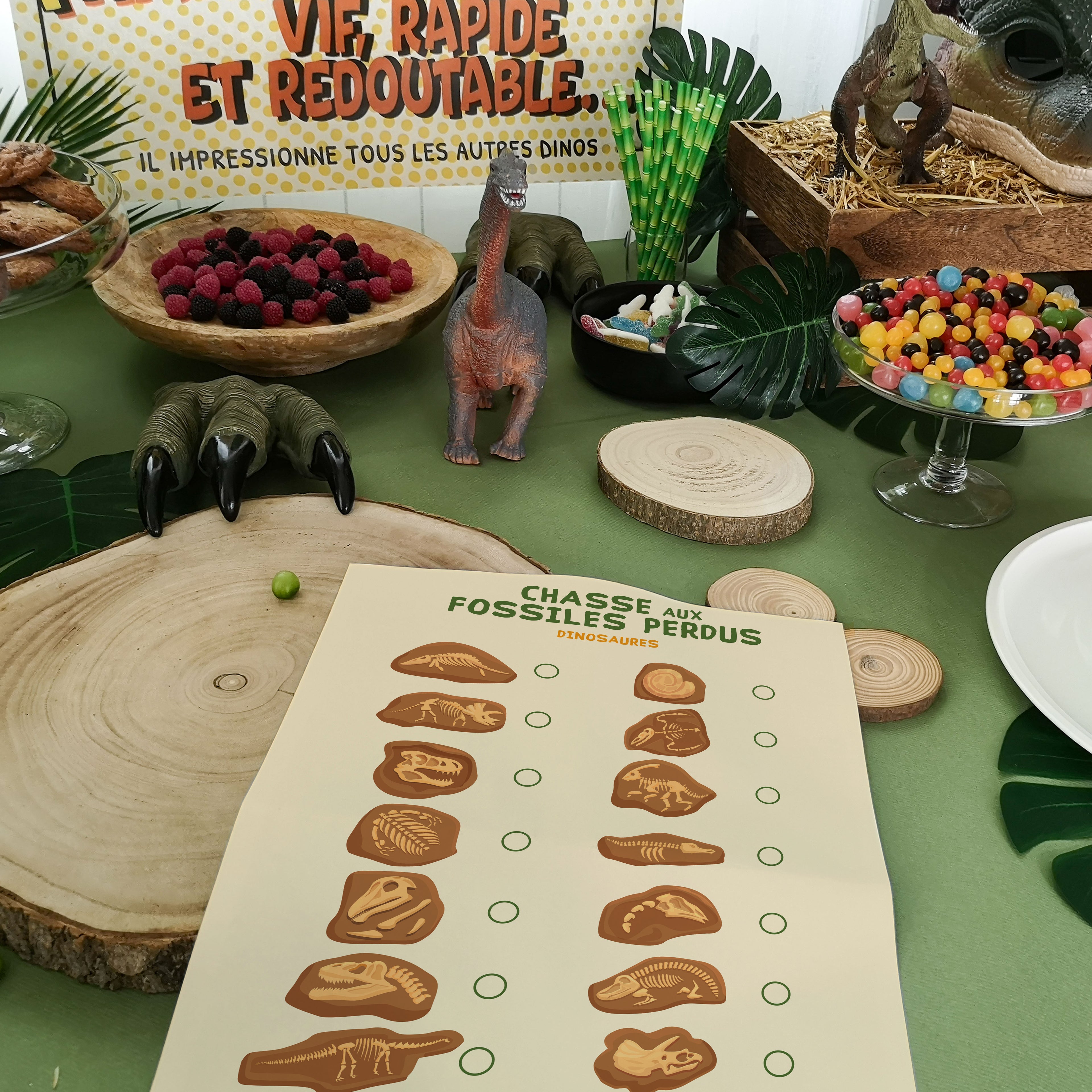 Animation de chasse aux fossiles sur le thème des dinosaures pour un anniversaire. La table décorée présente une fiche de jeu "Chasse aux fossiles perdus", des griffes de dino, des figurines, bonbons et décors tropicaux. Activité ludique pour enfants passionnés de paléontologie.
