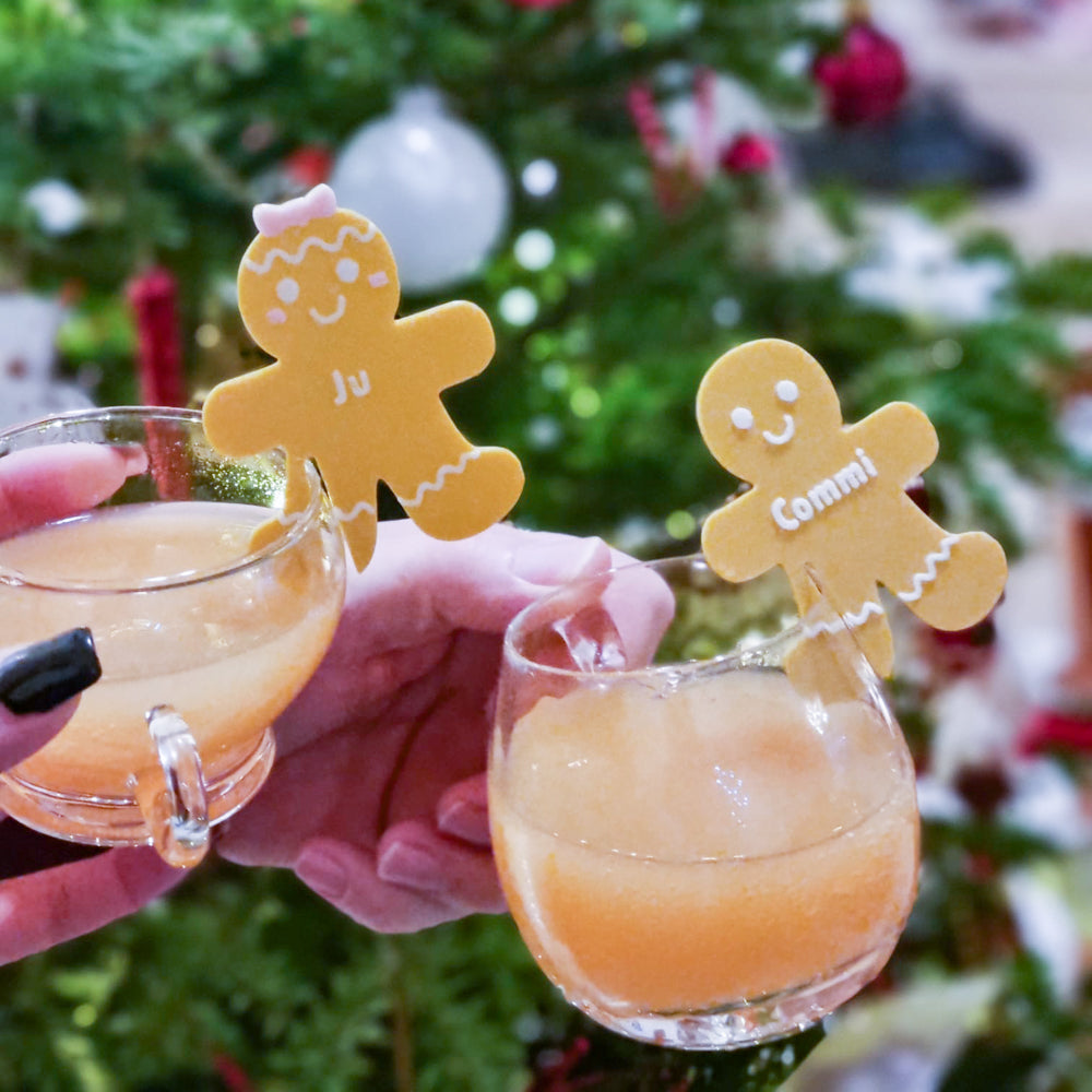 Deux mains trinquent devant un sapin de Noël, chaque verre décoré d’un marque-verre Bonhomme de pain d’épices personnalisé, apportant une touche chaleureuse et ludique aux fêtes de fin d’année.