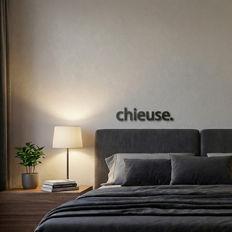 Mise en scène d’une décoration murale “chieuse.” en impression 3D (PLA) noire, fixée au-dessus d’un lit gris dans une chambre cosy, avec lampe de chevet allumée et petite plante sur une table de nuit.