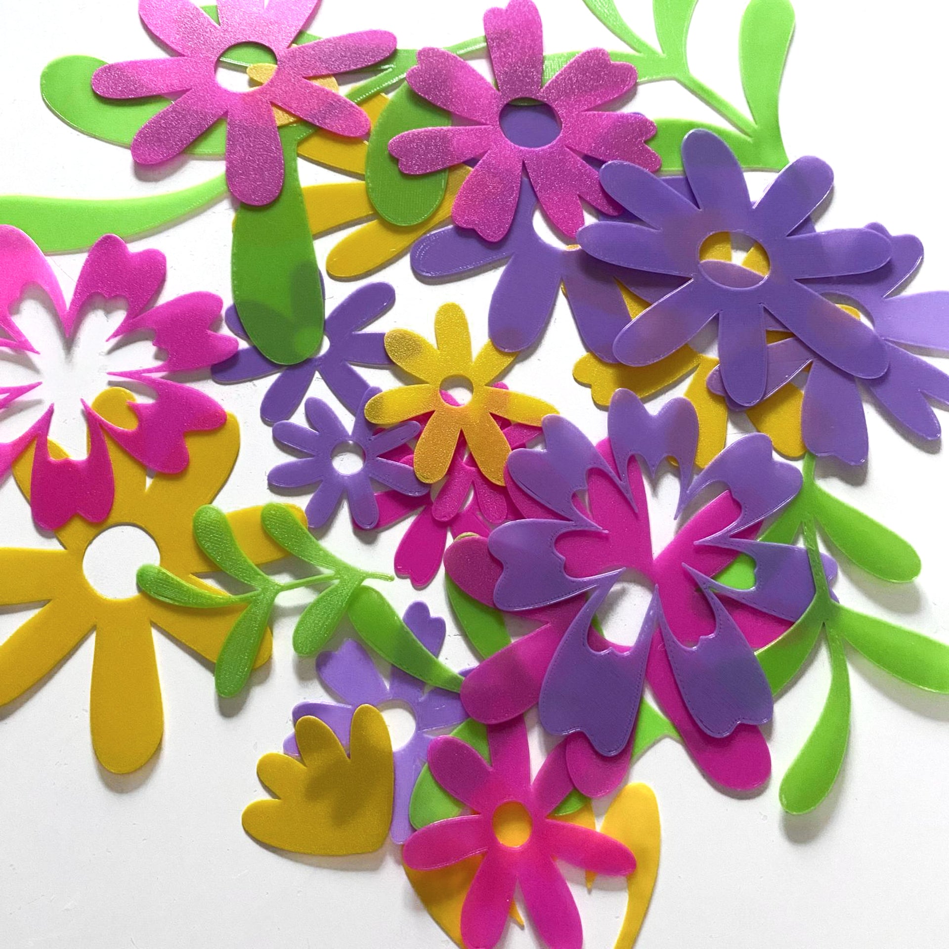 Assortiment de confettis de table en impression 3D PLA biodégradable "Magie Florale". Des formes de fleurs stylisées et de feuillages colorés en rose, magenta, violet, jaune et vert pomme sont éparpillés sur un fond blanc uni.