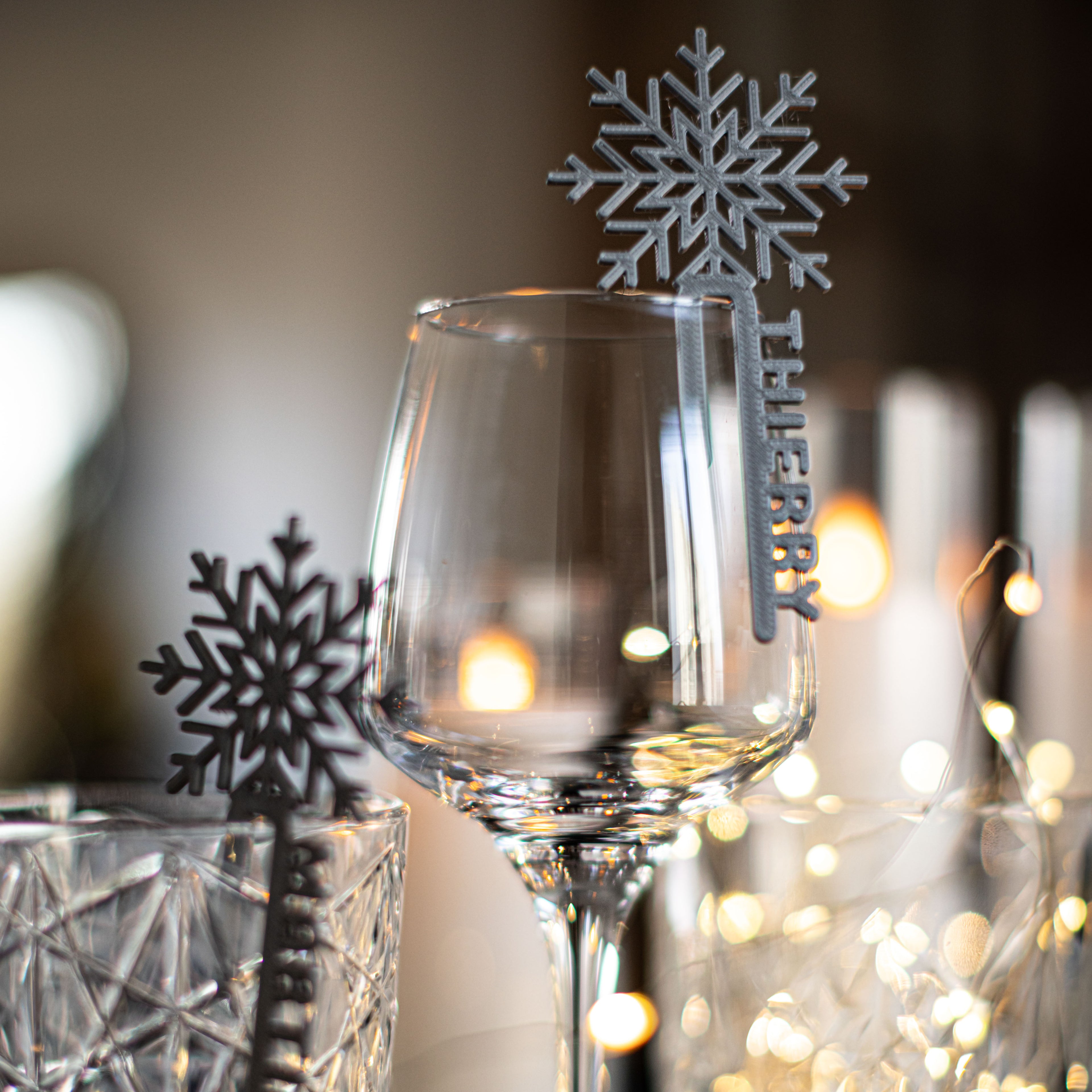 Marque-verre Flocon argenté personnalisé “Thierry” accroché à une coupe à vin, entouré de guirlandes lumineuses et de reflets chaleureux pour une ambiance hivernale élégante.