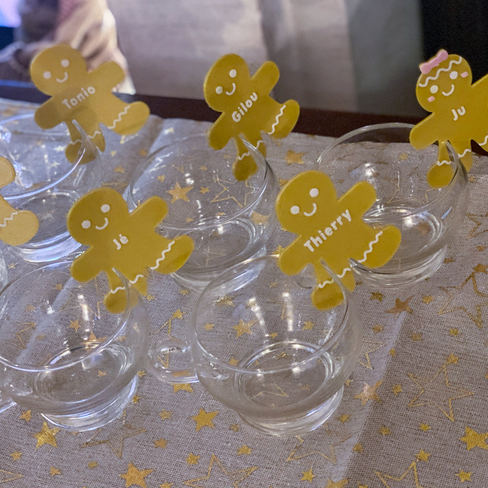 Alignement de verres transparents décorés de marque-verres Bonhomme de pain d’épices personnalisés (Tonio, Gilou, Ju, Jé, Thierry), posés sur une nappe étoilée dorée, en ambiance de préparation d’une table de Noël chaleureuse et festive.