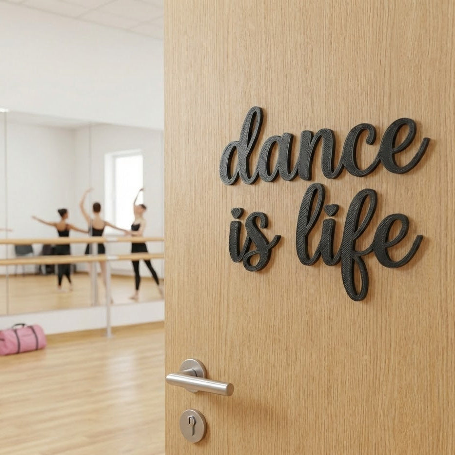 Décoration murale “dance is life” en impression 3D (PLA) noire, fixée sur une porte en bois, avec une salle de danse en arrière-plan (barres et danseuses floutées).