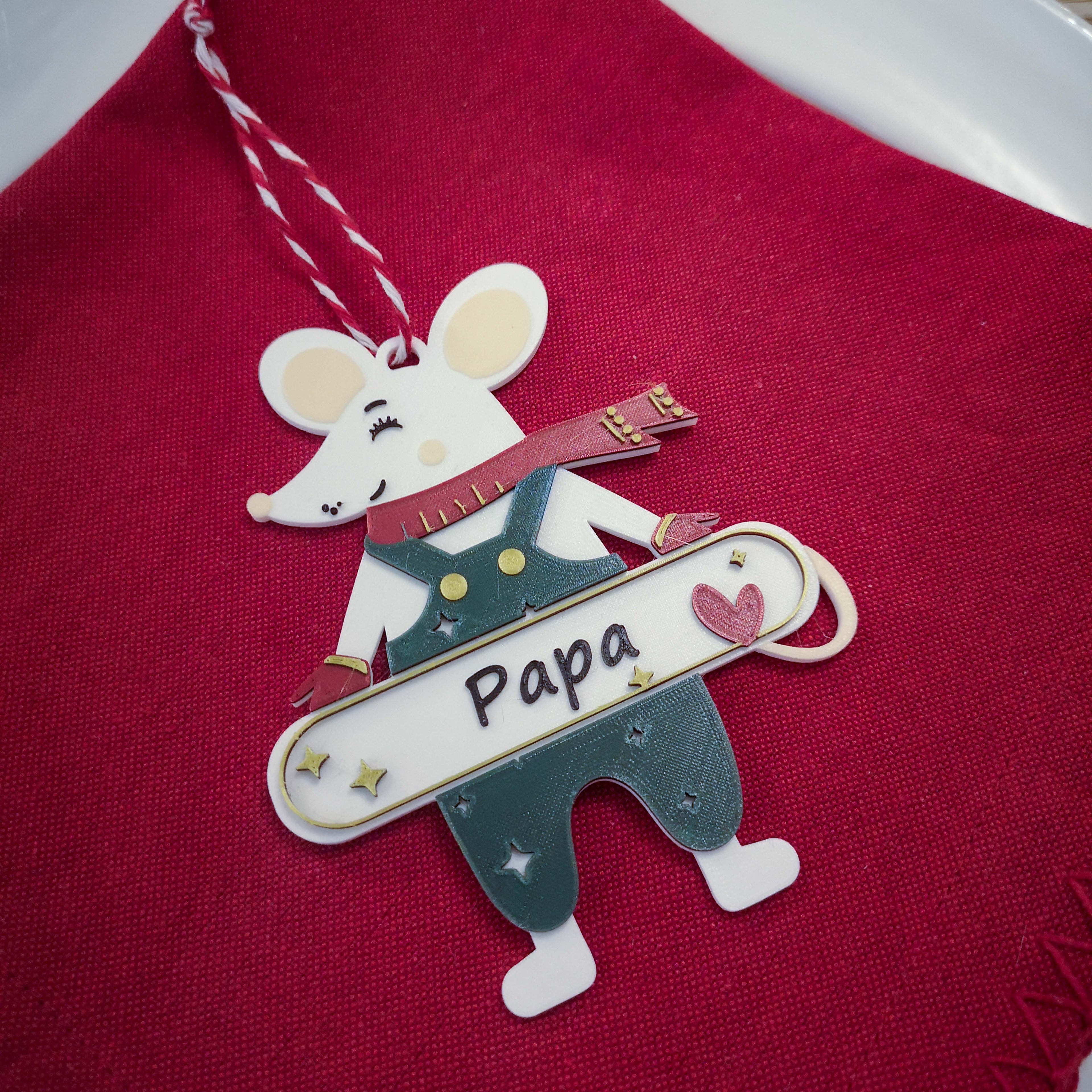 Gros plan d’une décoration de Noël personnalisable en forme de souris : souris blanche avec écharpe rouge et salopette, tenant une pancarte “Papa” avec un cœur, posée sur un tissu rouge, avec ficelle de suspension.
