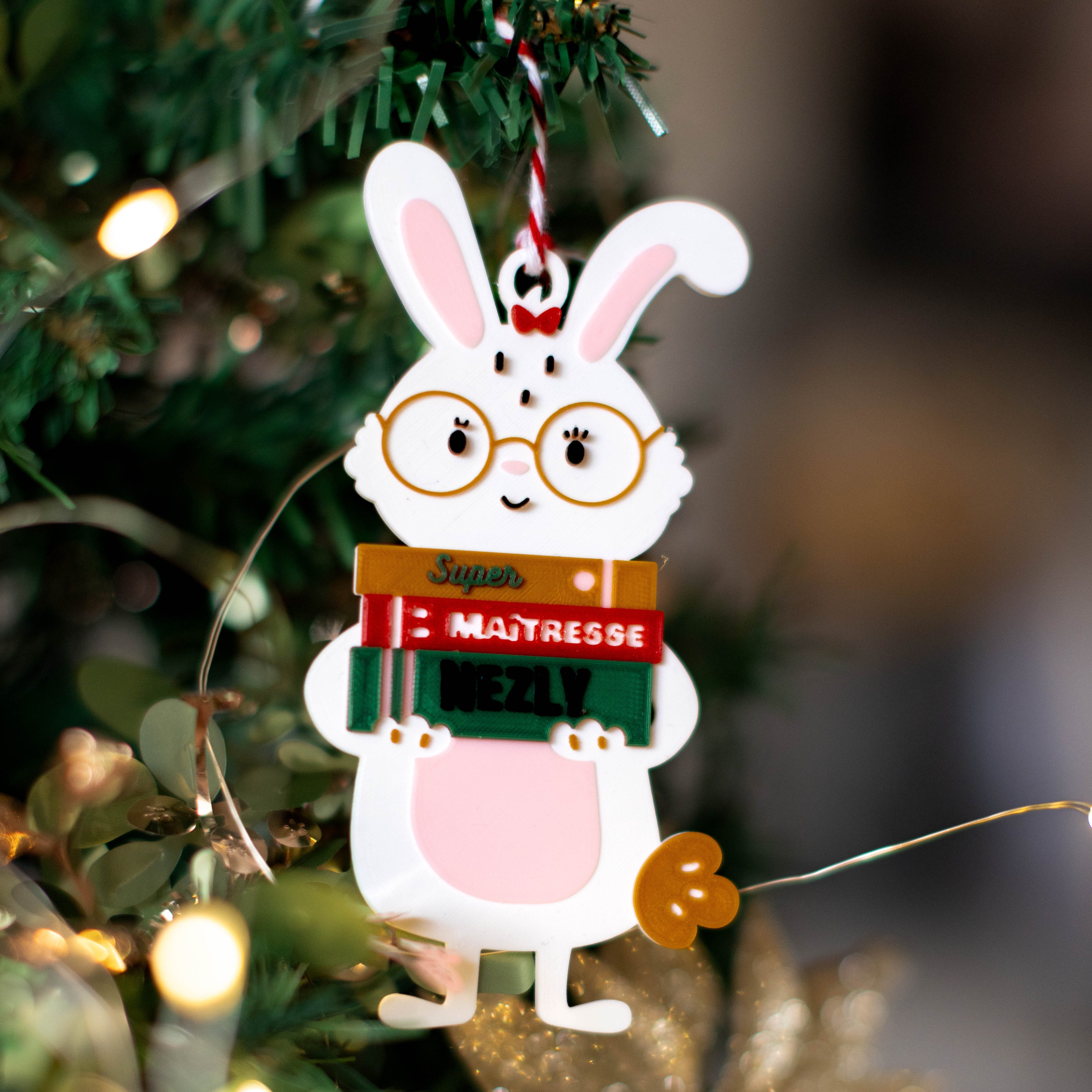 Décoration à suspendre sur sapin : lapin blanc avec lunettes, tenant des livres “Super”, “Maîtresse” et un prénom personnalisé (ex. “Nezly”), accroché à une branche de sapin avec guirlande lumineuse en arrière-plan.