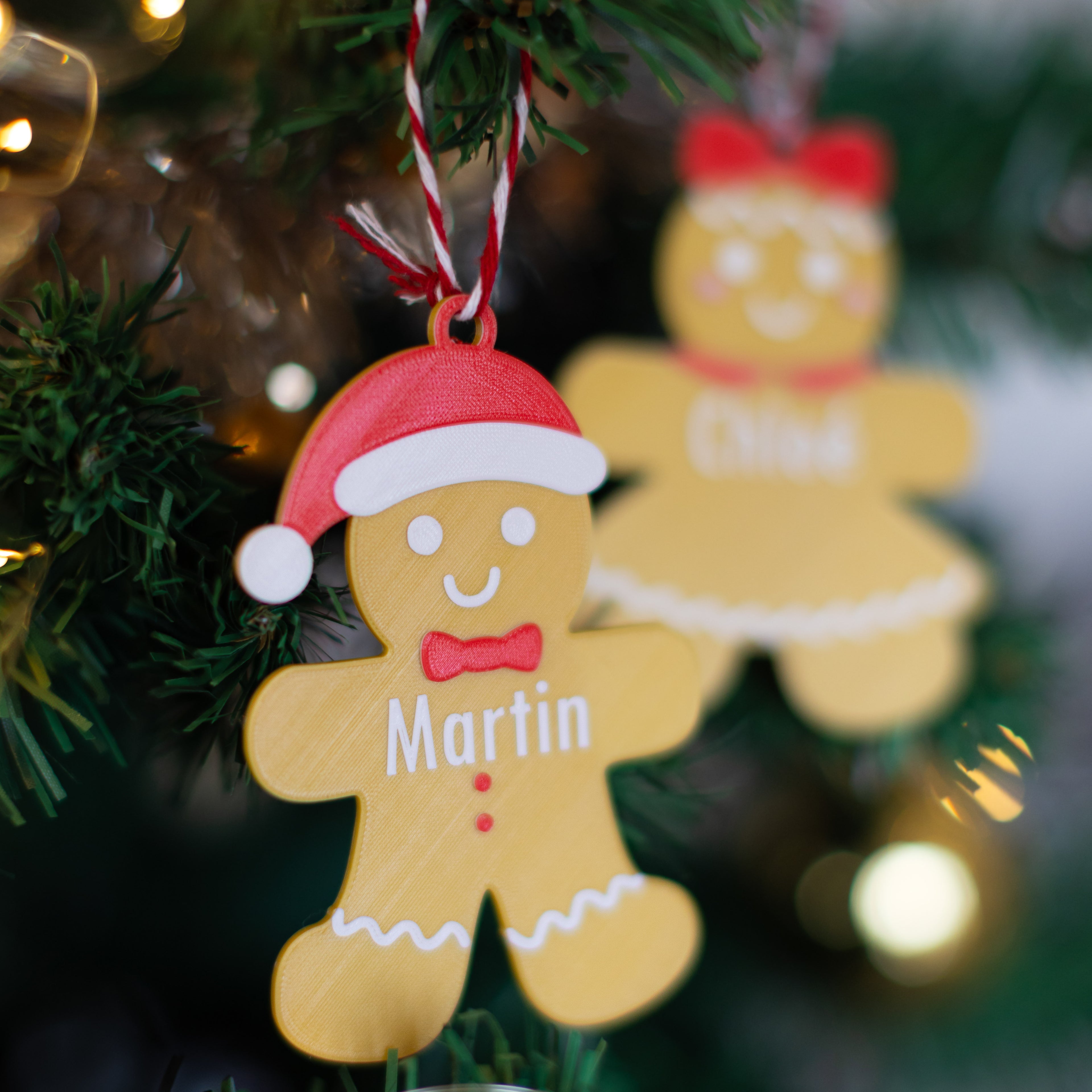 Bonhomme en pain d’épice personnalisable suspendu sur un sapin : décoration avec bonnet rouge de Père Noël, nœud papillon et prénom “Martin”, avec une seconde figurine floutée en arrière-plan.