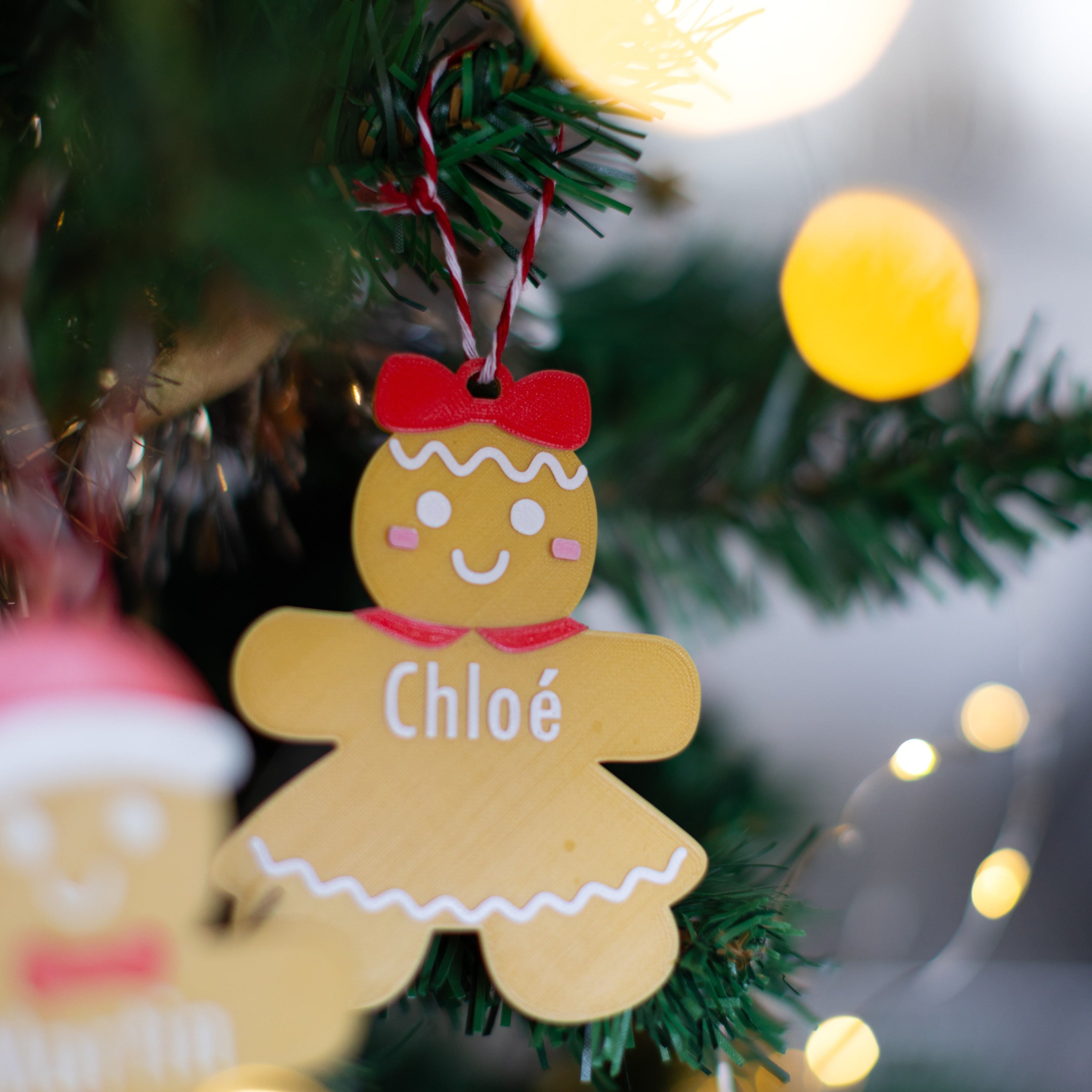Bonhomme en pain d’épice personnalisable suspendu sur un sapin : figurine avec nœud rouge, joues roses et prénom “Chloé”, sur fond de branches de sapin et lumières de Noël.