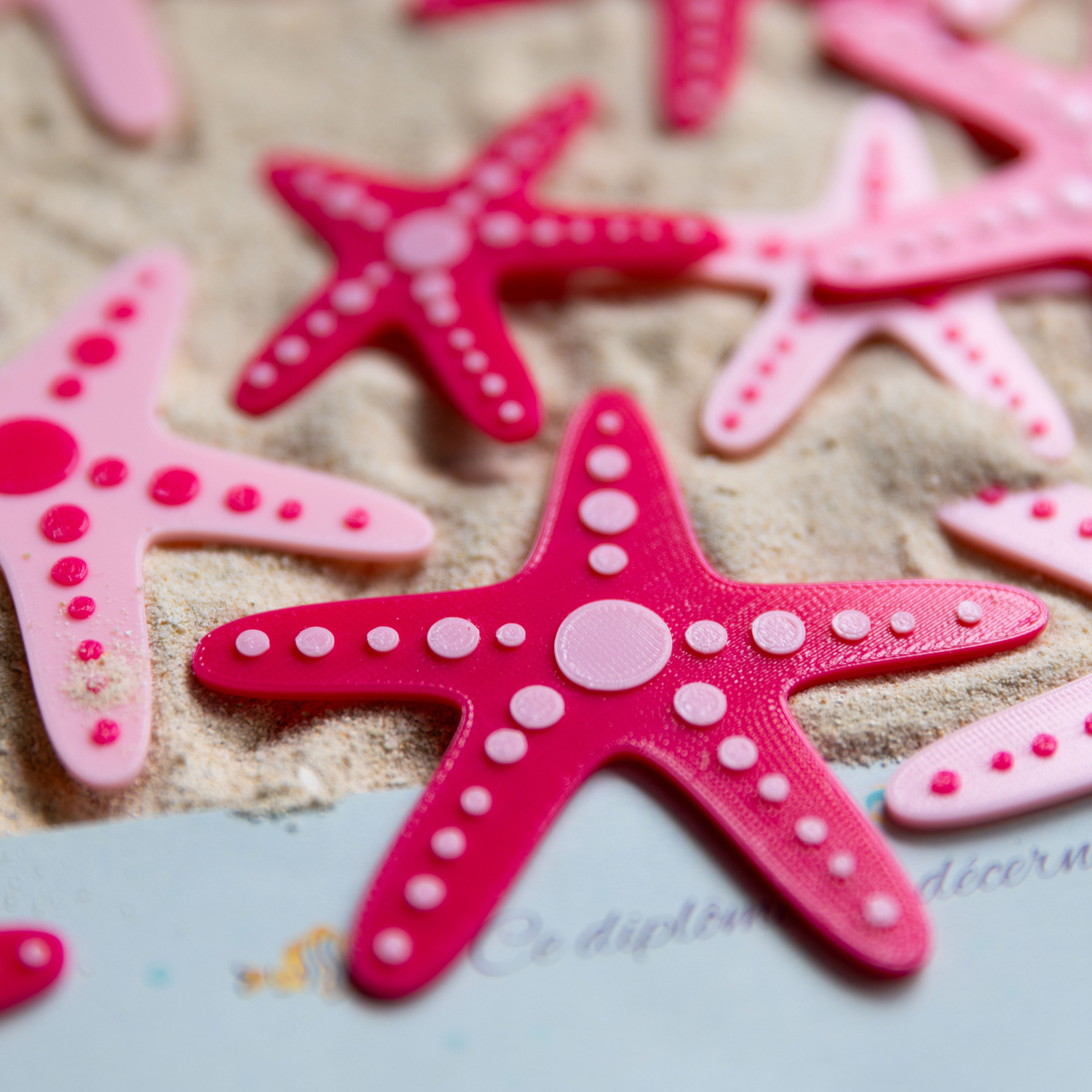 Deux modèles d’étoiles de mer bicolores (rose clair et fuchsia) réalisées en impression 3D, disposées sur du sable pour une activité de chasse au trésor sur le thème des sirènes.