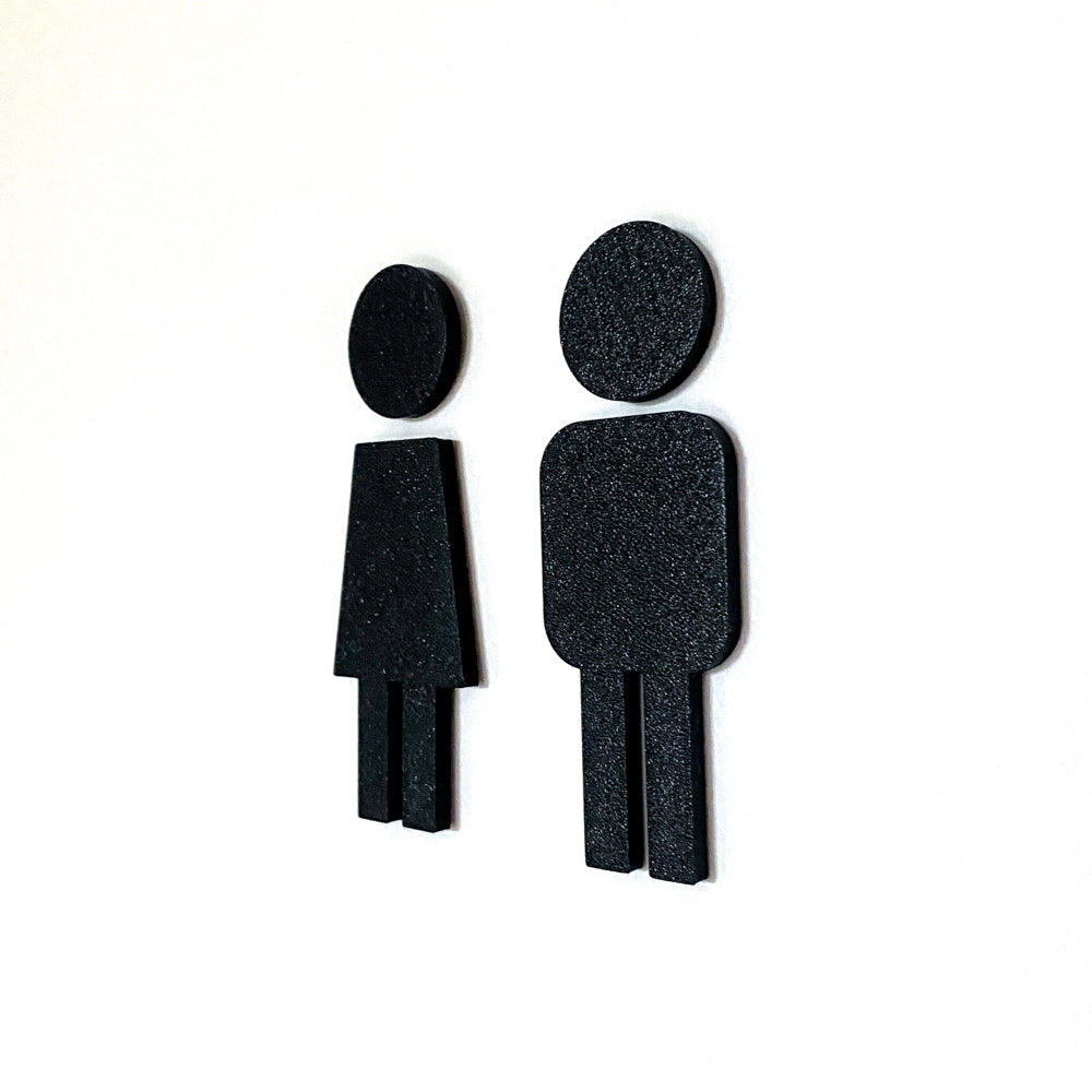 Gros plan sur deux pictogrammes noirs fille et garçon en relief, collés sur un fond blanc pour signalétique de toilettes familiales ou WC mixtes.
