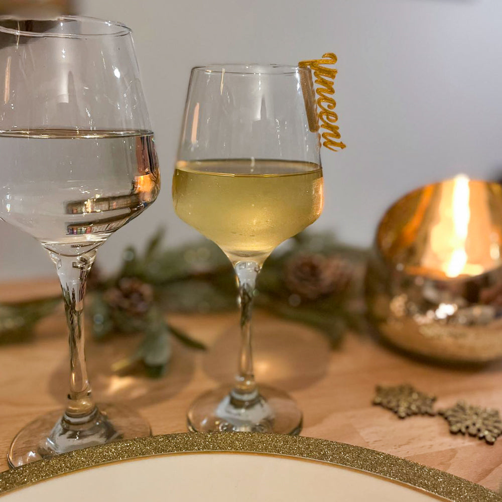 Deux verres à pied posés sur une table décorée : l’un rempli d’eau, l’autre de vin blanc, avec sur ce dernier un marque-verre doré en écriture cursive portant le prénom “Vincent”, clipsé au bord du verre. En arrière-plan, une décoration de fête avec branches, pommes de pin et une bougie dorée diffuse une lumière chaleureuse.