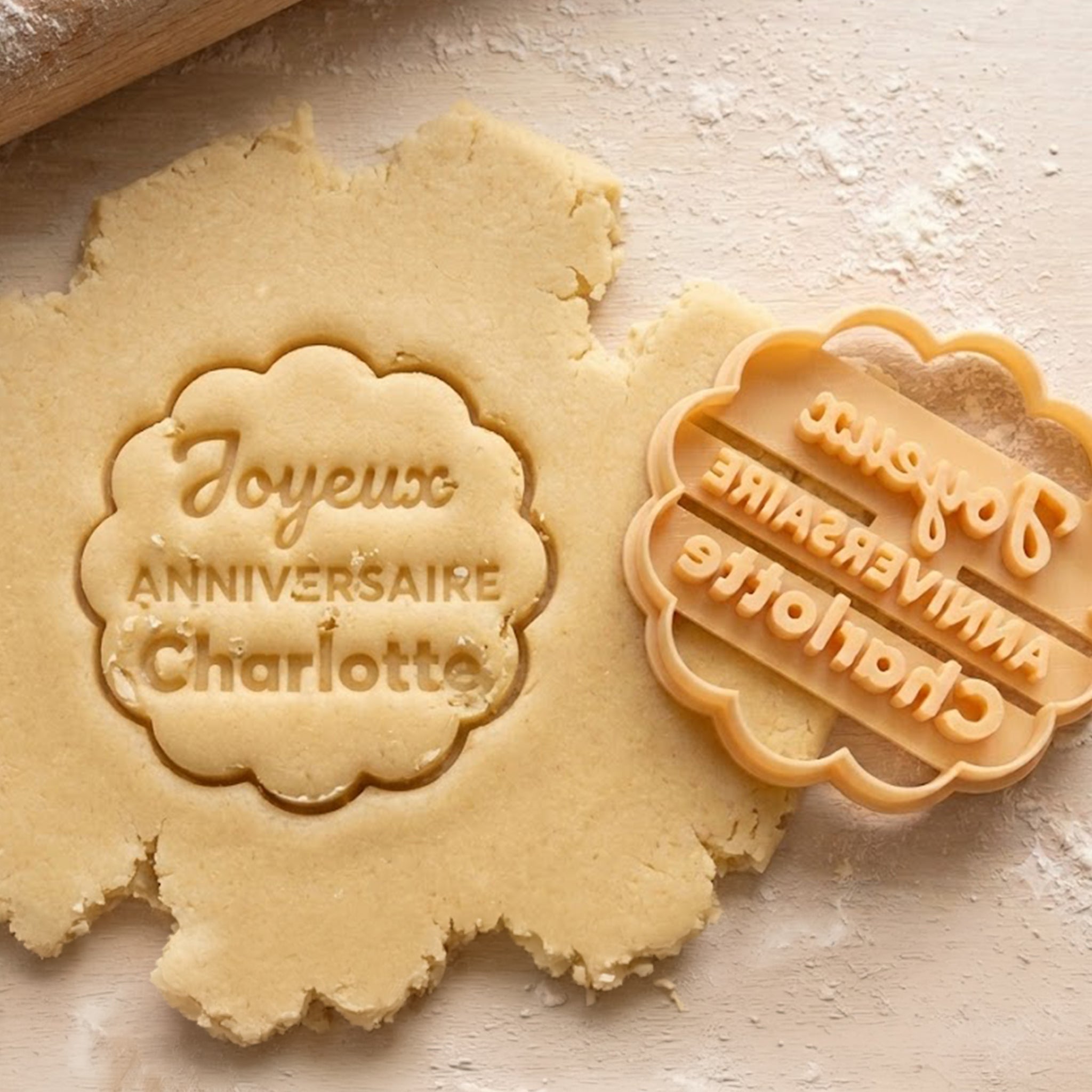 Gros plan sur la pâte à biscuit crue avec l'empreinte gravée "Joyeux ANNIVERSAIRE Charlotte" et l'emporte-pièce à côté, avec un rouleau à pâtisserie visible sur une table farinée.