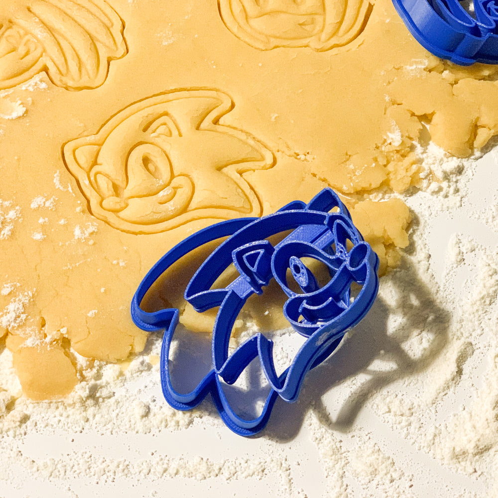 Emporte-pièces Turbo Hérisson créant des empreintes de biscuits représentant Sonic, Tails et Knuckles dans une pâte sablée, avec les moules bleus visibles autour de la pâte.