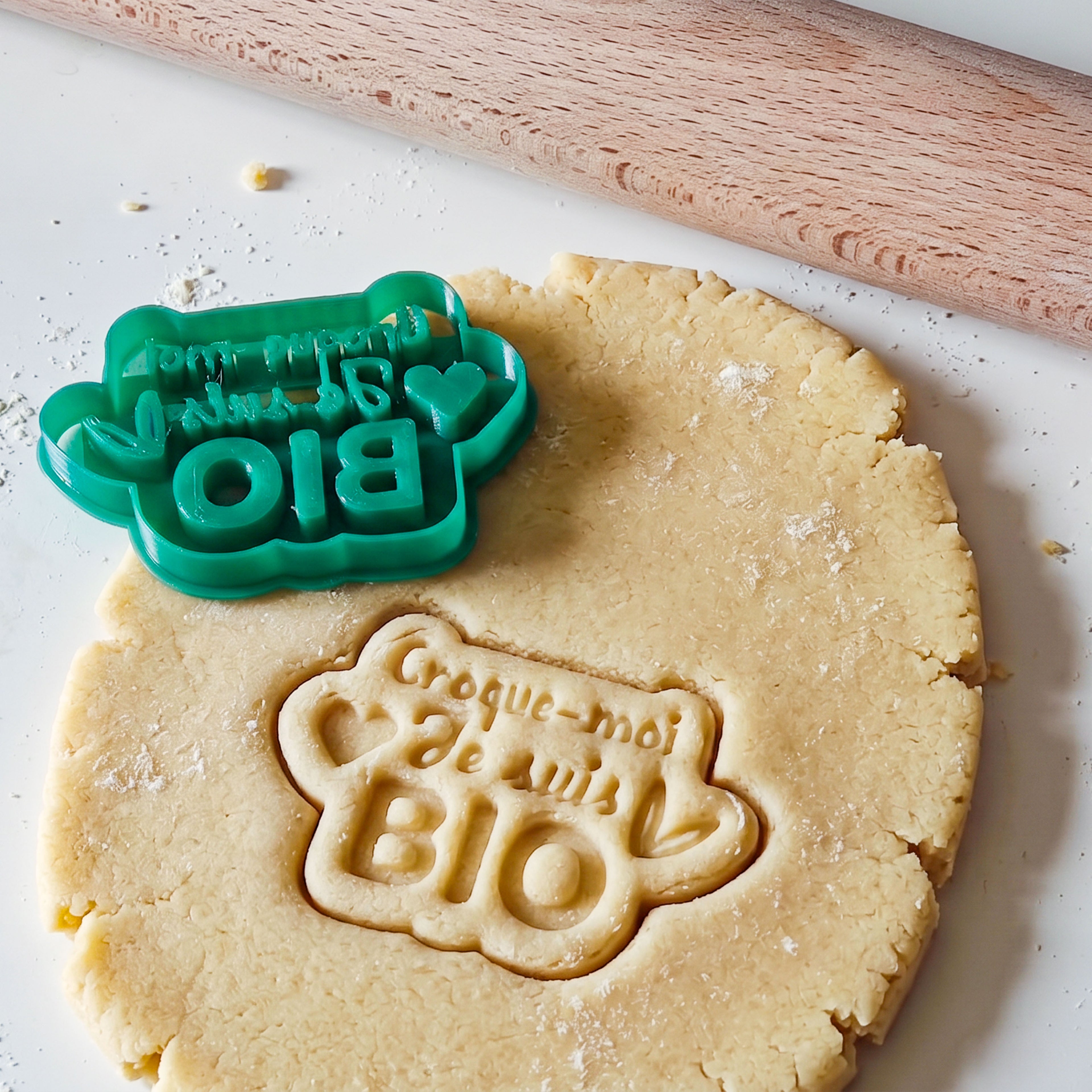Photographie détaillée montrant un emporte-pièce personnalisé vert imprimé en 3D en cours d'utilisation sur de la pâte à biscuit étalée. L'emporte-pièce laisse une impression nette et lisible du texte 'Croque-moi je suis BIO' avec des motifs de cœur et de plante stylisés. Un coin de rouleau à pâtisserie en bois est visible en arrière-plan sur une surface farinée