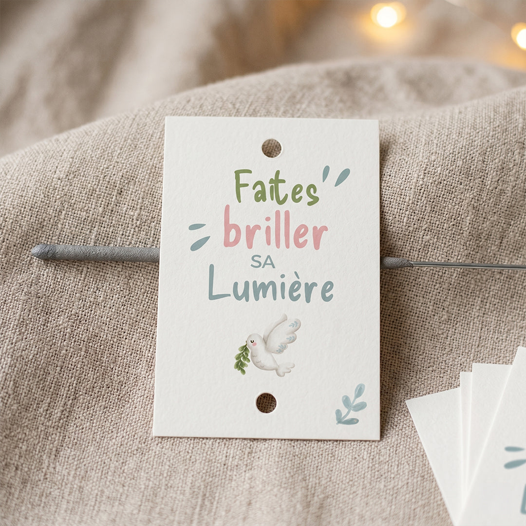 Gros plan sur une étiquette blanche perforée pour baptême ou communion avec l'inscription "Faites' briller SA Lumière" et un dessin de colombe, posée sur un tissu texturé beige.