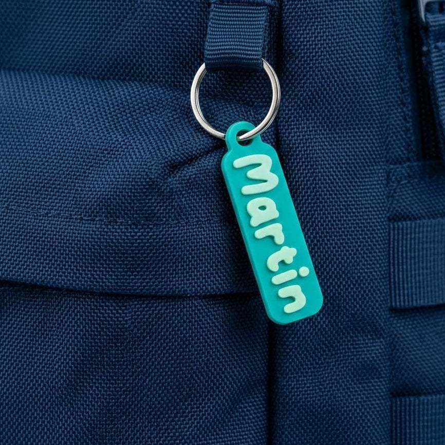Étiquette 3D prénom « Martin » en PLA turquoise avec lettres gris-bleues, sur anneau métal, accrochée à un sac bleu.