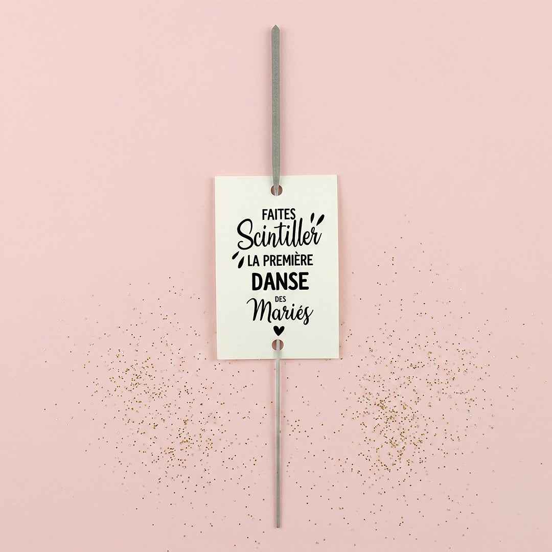 Gros plan d'une étiquette rectangulaire blanche pour cierge magique, affichant le texte "FAITES SCINTILLER LA PREMIÈRE DANSE DES MARIÉS" avec un petit cœur. Un cierge magique est inséré au centre. Fond rose avec des paillettes dorées éparpillées.