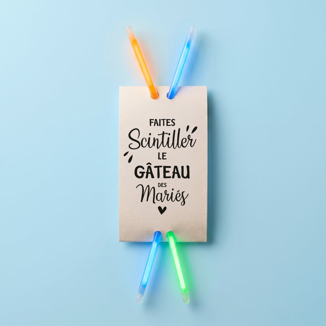 Vue de dessus d'une étiquette rectangulaire verticale blanche avec l'inscription "FAITES Scintiller LE GÂTEAU DES Mariés". Des bracelets fluo lumineux colorés sont glissés à travers les perforations, le tout sur un fond bleu uni.