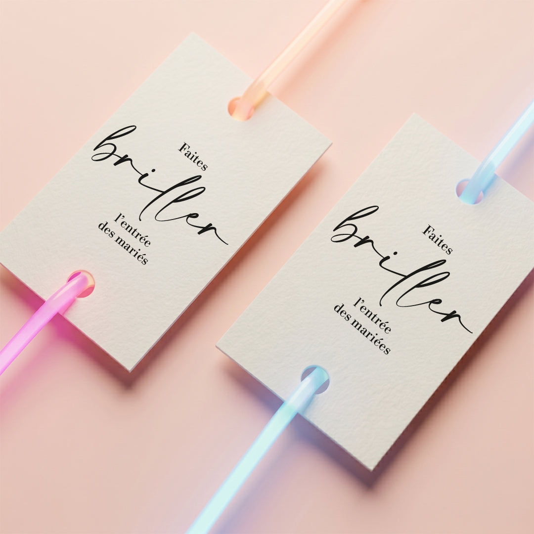 Vue de dessus carrée de deux étiquettes de mariage blanches au format 50x73mm sur un fond rose pastel uni et lumineux. L'étiquette de gauche affiche le texte "Faites briller l'entrée des mariés" et celle de droite "Faites briller l'entrée des mariées". Un bracelet néon rose/orange et un bleu lumineux sont enfilés à travers deux trous et passent proprement derrière chaque étiquette.