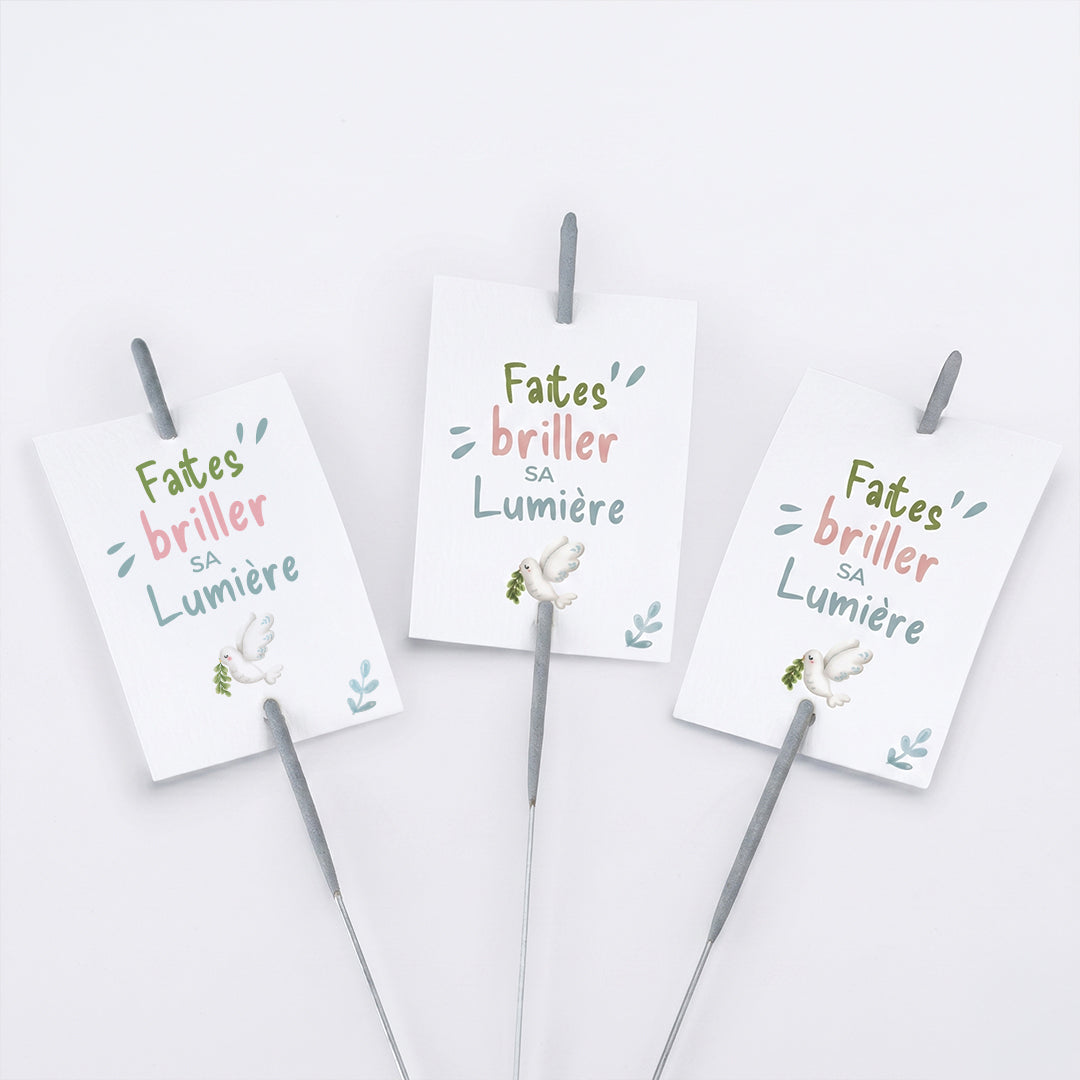 Trois étiquettes rectangulaires blanches pour baptême ou communion avec l'inscription "Faites' briller SA Lumière" et une illustration de colombe. Un cierge magique est glissé verticalement dans chaque étiquette, présentées sur un fond blanc.