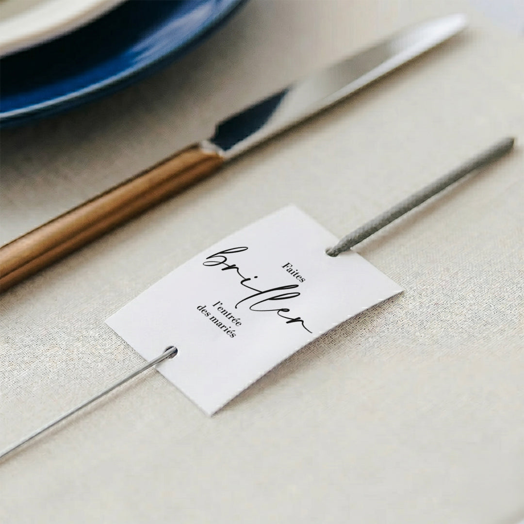 Gros plan sur une étiquette rectangulaire en papier blanc, percée de deux trous dans lesquels passe la tige grise d'un cierge magique. L'étiquette comporte l'inscription en français « Faites briller l'entrée des mariés » avec une écriture cursive élégante pour le mot « briller ». L'ensemble repose sur une nappe en lin beige clair. En arrière-plan flou, on aperçoit une assiette bleu foncé et un couteau de table au manche doré.