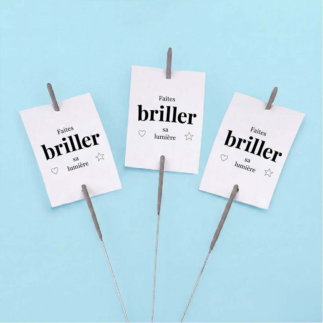 Trois étiquettes rectangulaires blanches "Faites briller sa lumière" avec un petit cœur et une étoile. Un cierge magique est glissé verticalement dans chaque étiquette, présentées sur un fond bleu clair.