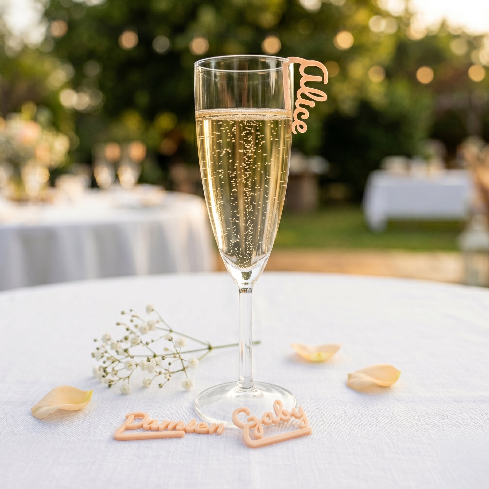Flûte remplie de champagne lors d'un mariage estival en extérieur. Un marque-verre personnalisé de couleur Abricot Pastel avec le prénom Alice est accroché au rebord du verre. Au pied du verre, sur une nappe en lin blanc, deux autres marque-places aux prénoms Damien et Gaby sont posés à côté d'un brin de gypsophile et de pétales de rose. L'arrière-plan lumineux et flou dévoile une ambiance festive de réception avec des guirlandes lumineuses de guinguette.