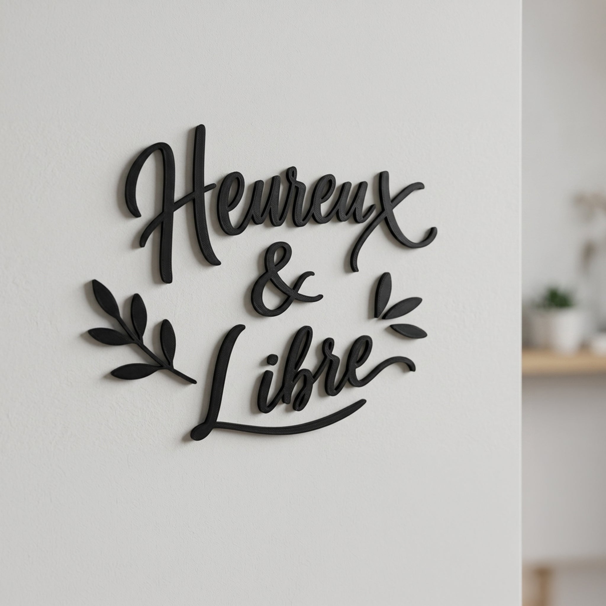 Décoration murale en impression 3D noire affichant le texte calligraphié « Heureux & Libre » sur trois lignes avec des motifs de feuilles. Le lettrage en relief de 3 mm est fixé sur un mur blanc épuré et projette une ombre portée élégante.
