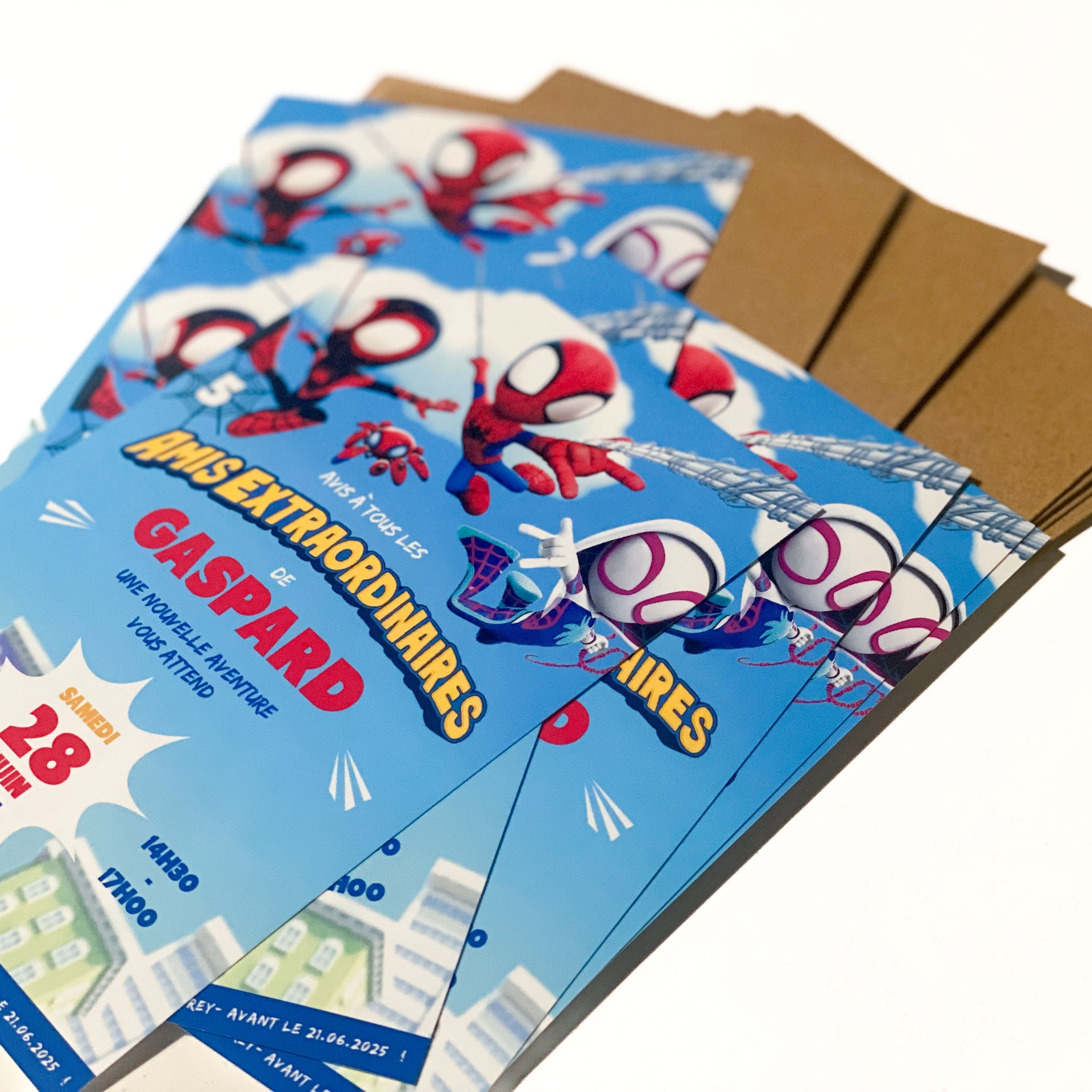 Lot d’invitations d’anniversaire posées en éventail sur des enveloppes kraft. Visuel bleu ciel façon BD : deux héros masqués rouges et blancs se balancent à des toiles, nuages et immeubles en arrière-plan. Texte lisible : “Amis extraordinaires”, prénom “GASPARD” en rouge, date “Samedi 28 juin”, horaires “14h30–17h00”