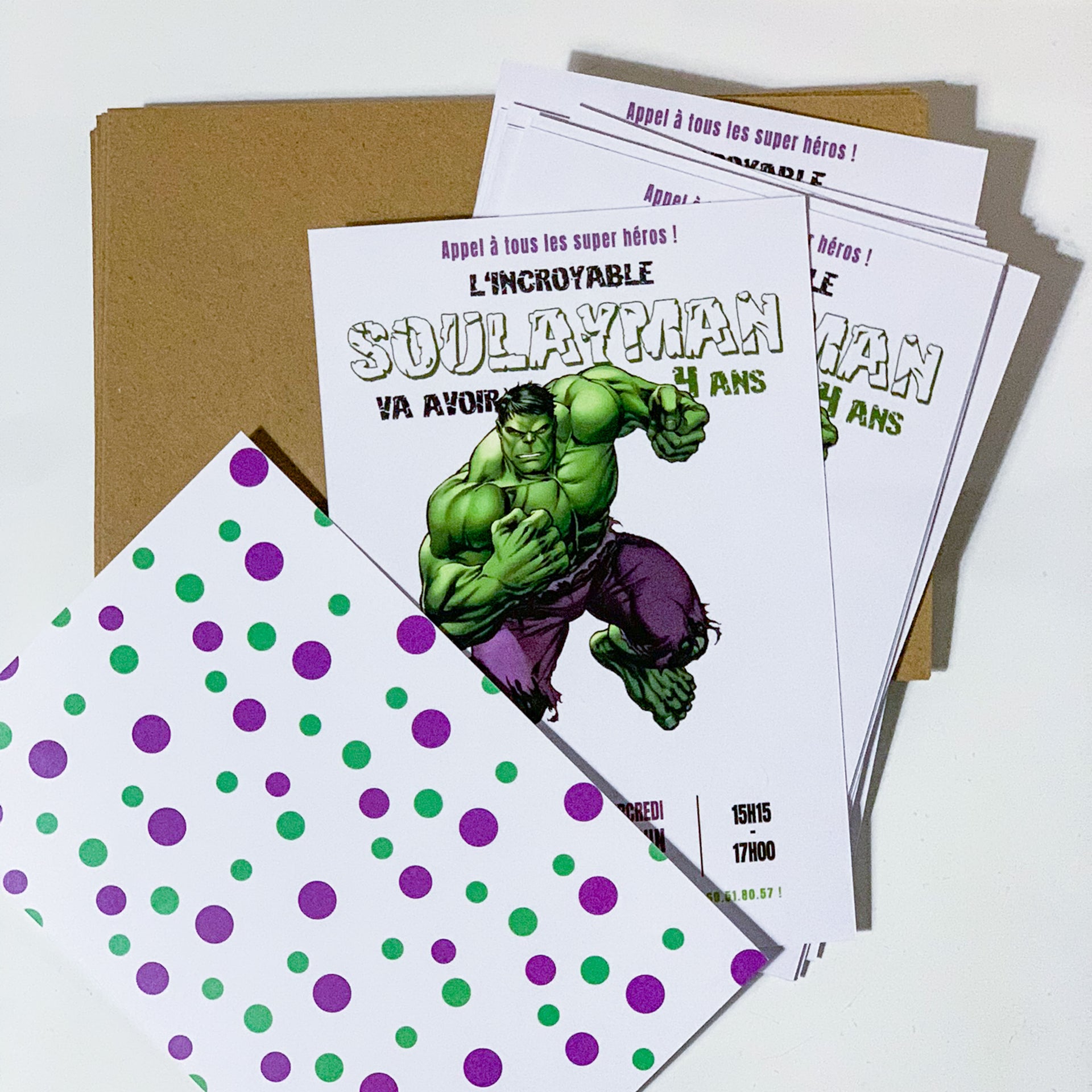 Empilement d’invitations d’anniversaire imprimées posées sur des enveloppes kraft. Au centre de chaque carte, un grand super-héros vert musclé en short violet, sur fond blanc. En haut, le titre “L’INCROYABLE Soulayman” en lettres épaisses, détails de fête en bas à droite. Un verso blanc à pois verts et violets est posée au premier plan.