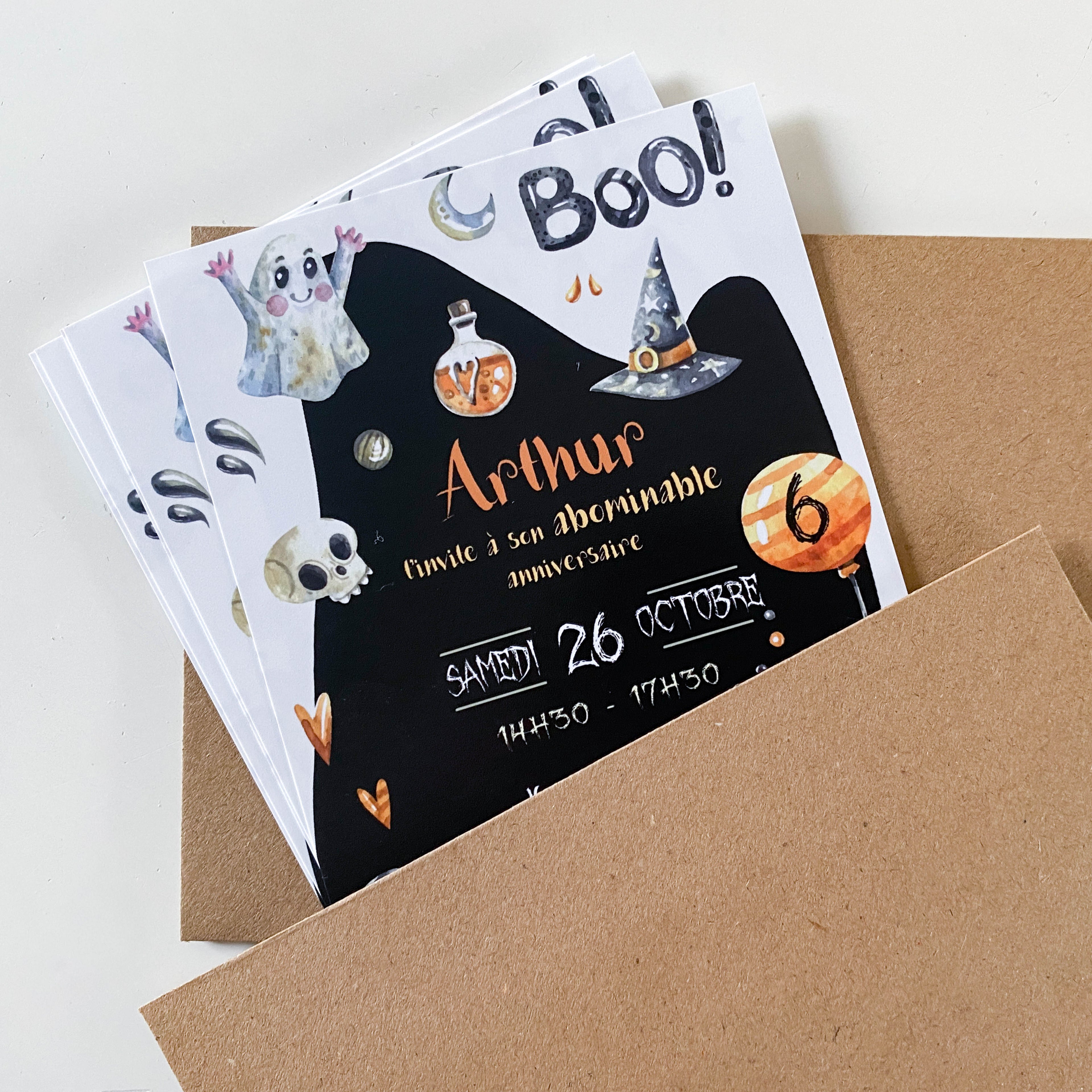Trois invitations d’anniversaire Halloween glissées dans des enveloppes kraft. Fond noir en forme de fantôme avec le titre “Boo !”. Illustrations aquarelle : chapeau de sorcière, fiole, lune, crâne et petites gouttes. Prénom “Arthur” en orange, date “Samedi 26 octobre”, horaires “14h30–17h30”, ballon orange portant le chiffre 6.