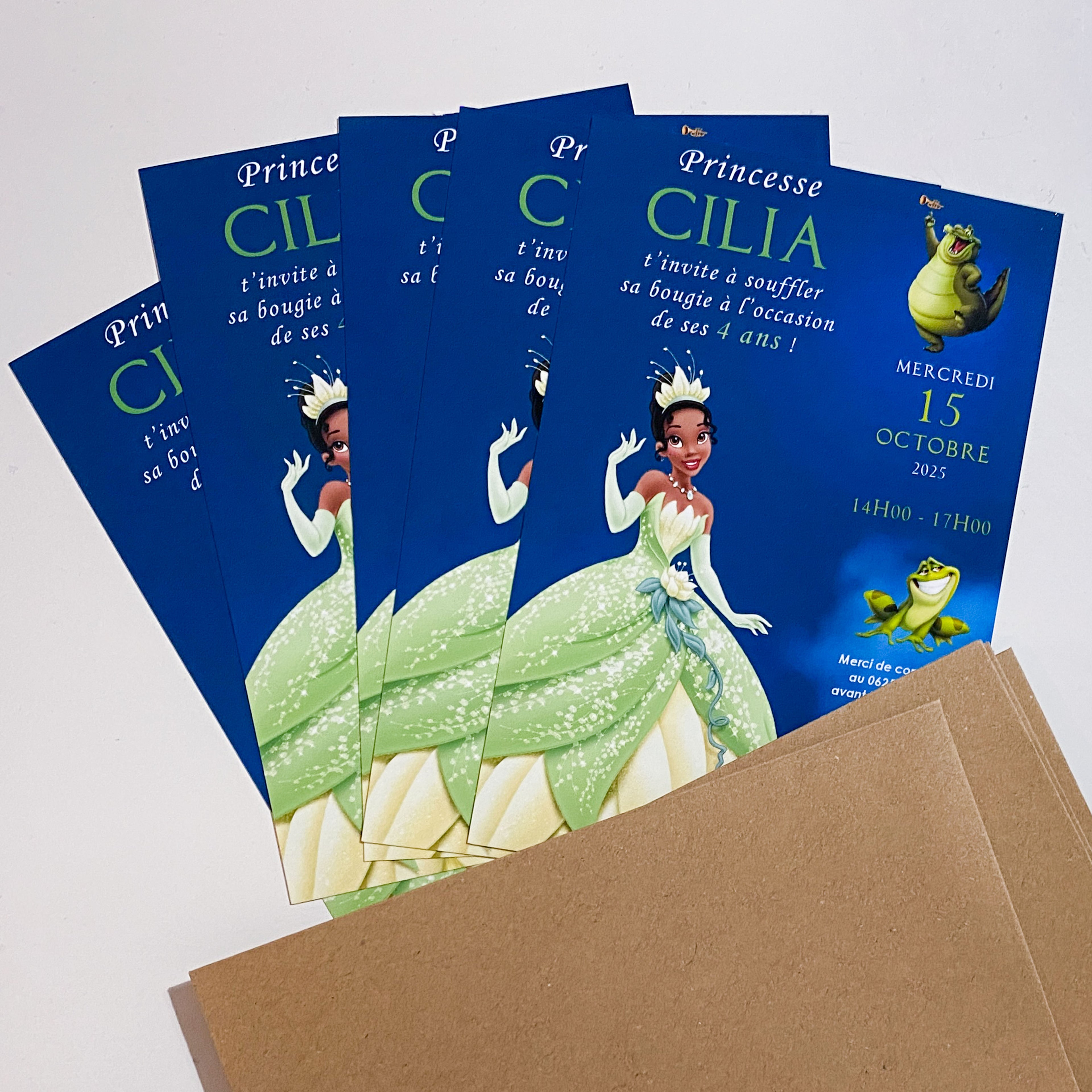Cinq invitations d’anniversaire imprimées  posées en éventail sur des enveloppes kraft. Fond bleu nuit, grande princesse brune souriante en robe de bal vert clair pailletée et diadème. À droite, un petit alligator et une grenouille. Gros prénom “CILIA” en haut, puis texte, date “15 octobre 2025” et horaires en colonnes.